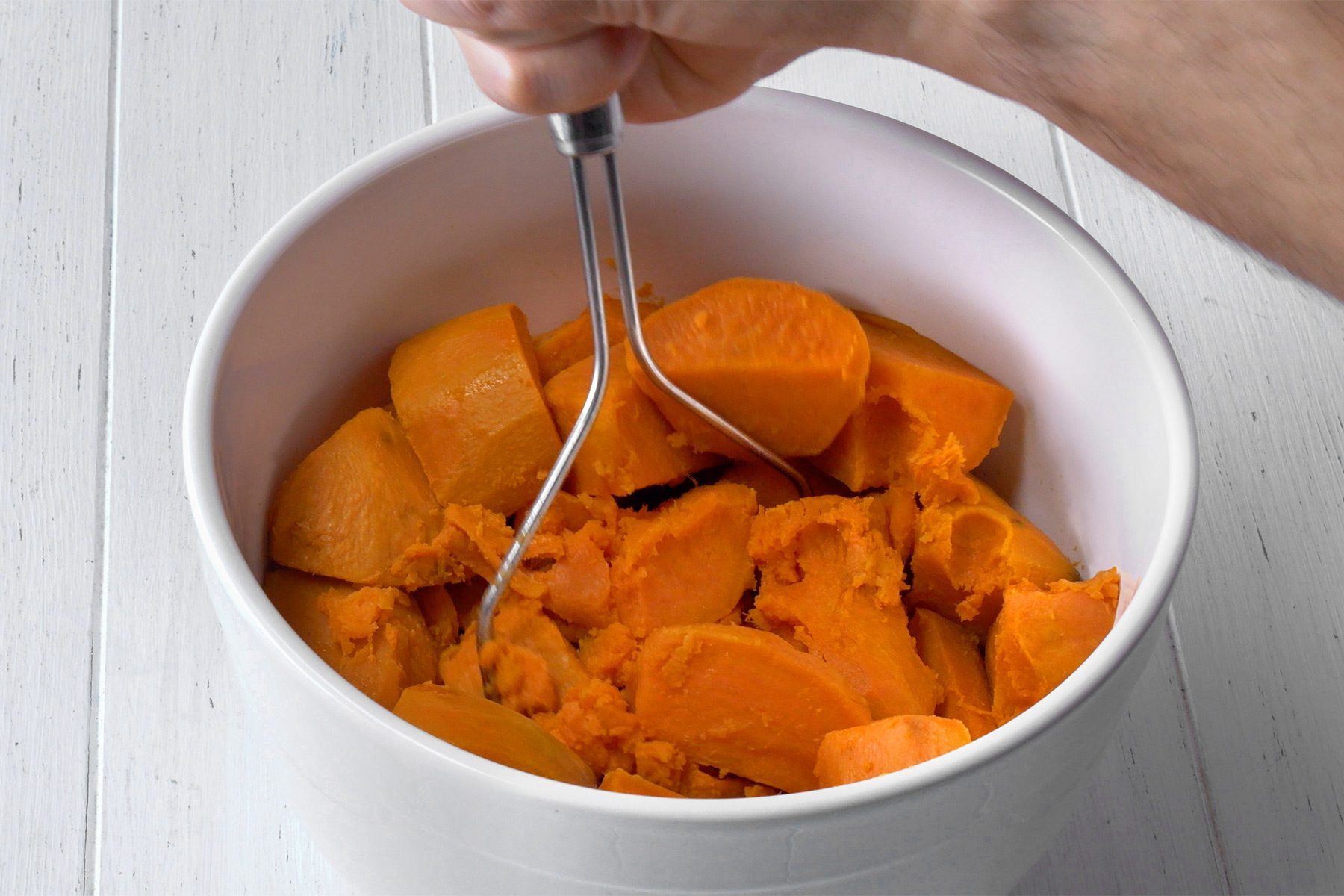 A close-up image of a person mashing chunks of cooked sweet potatoes in a white ceramic bowl using a stainless steel potato masher. The bowl is placed on a white wooden surface. The person’s hand gripping the masher is visible in the top right corner.