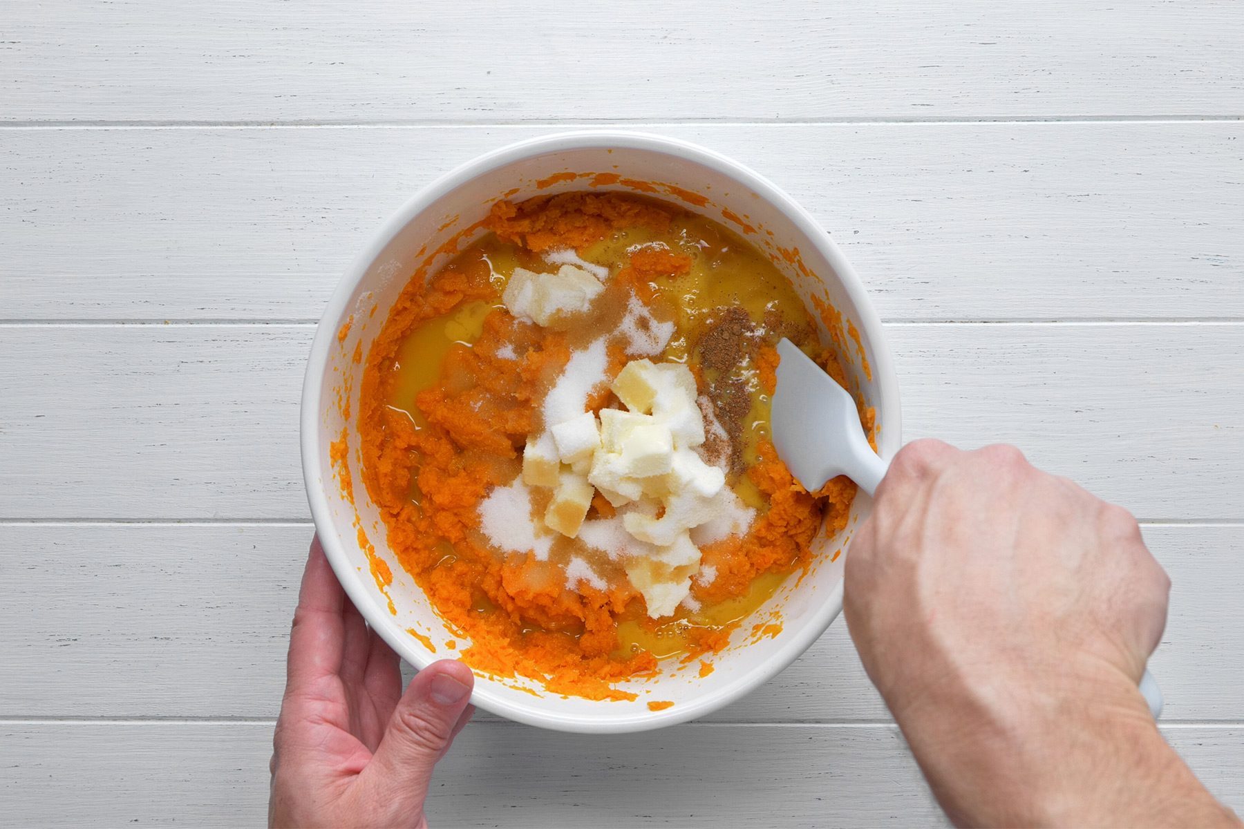 A person's hands are seen holding and mixing ingredients with a spatula in a white bowl. The bowl contains a mixture of mashed carrots, eggs, sugar, and other ingredients, likely being prepared for baking. The background is a light wooden surface.
