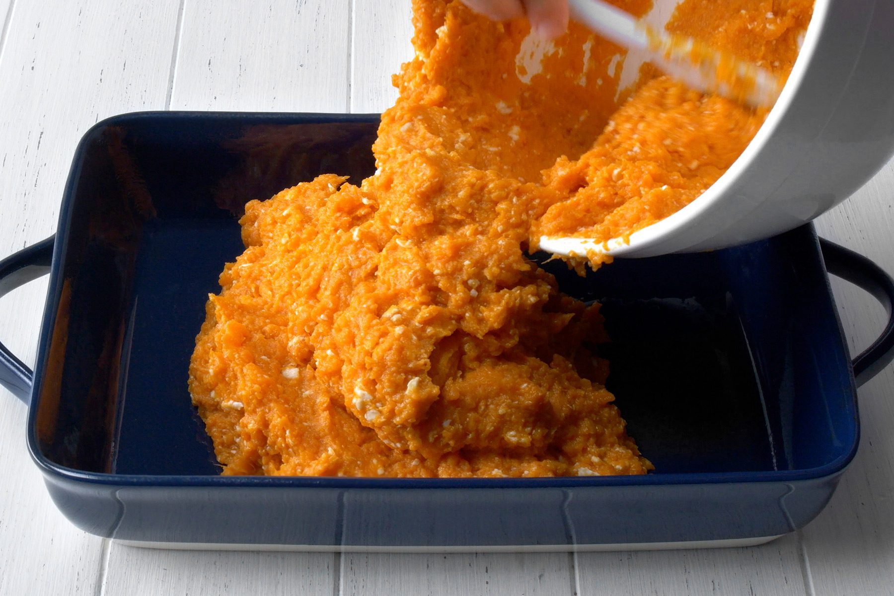 A person is pouring mashed sweet potatoes from a white bowl into a rectangular blue baking dish. The baking dish is placed on a white wooden surface.