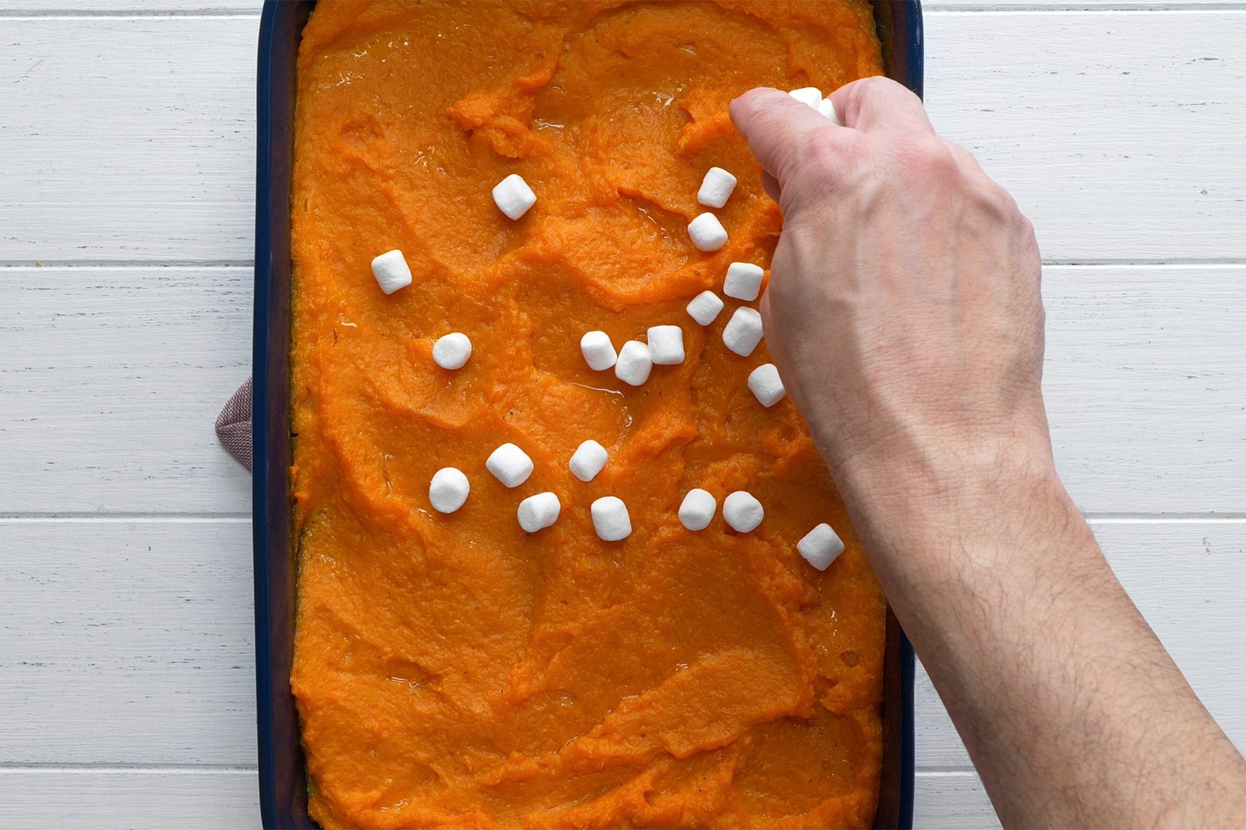 A person's hand sprinkles mini marshmallows on top of a smooth layer of mashed sweet potatoes in a rectangular baking dish. The dish rests on a white wood surface.