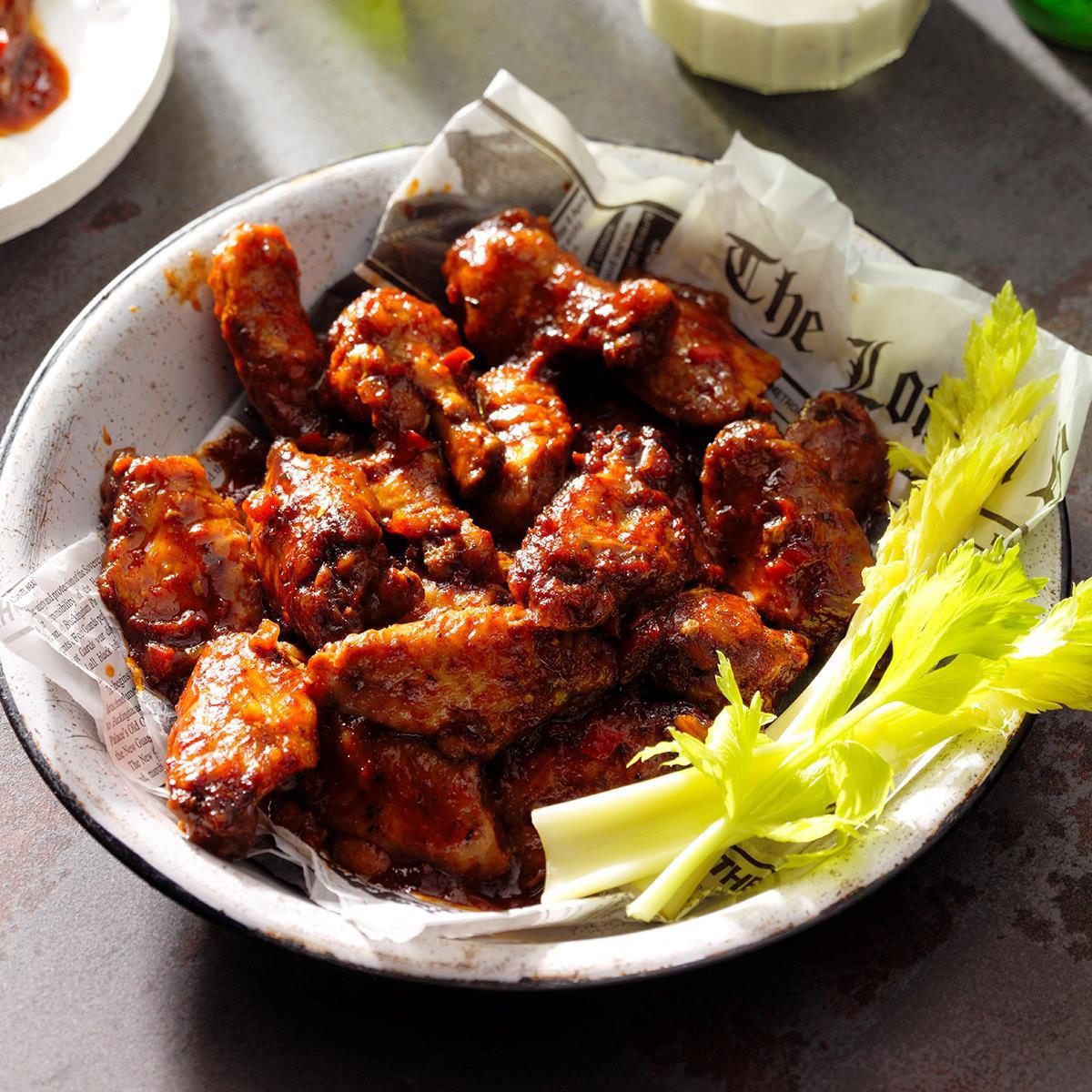 A bowl filled with succulent, glazed chicken wings garnished with fresh celery stalks is placed on a textured surface. The bowl is lined with a piece of newspaper, adding a rustic touch to the presentation.