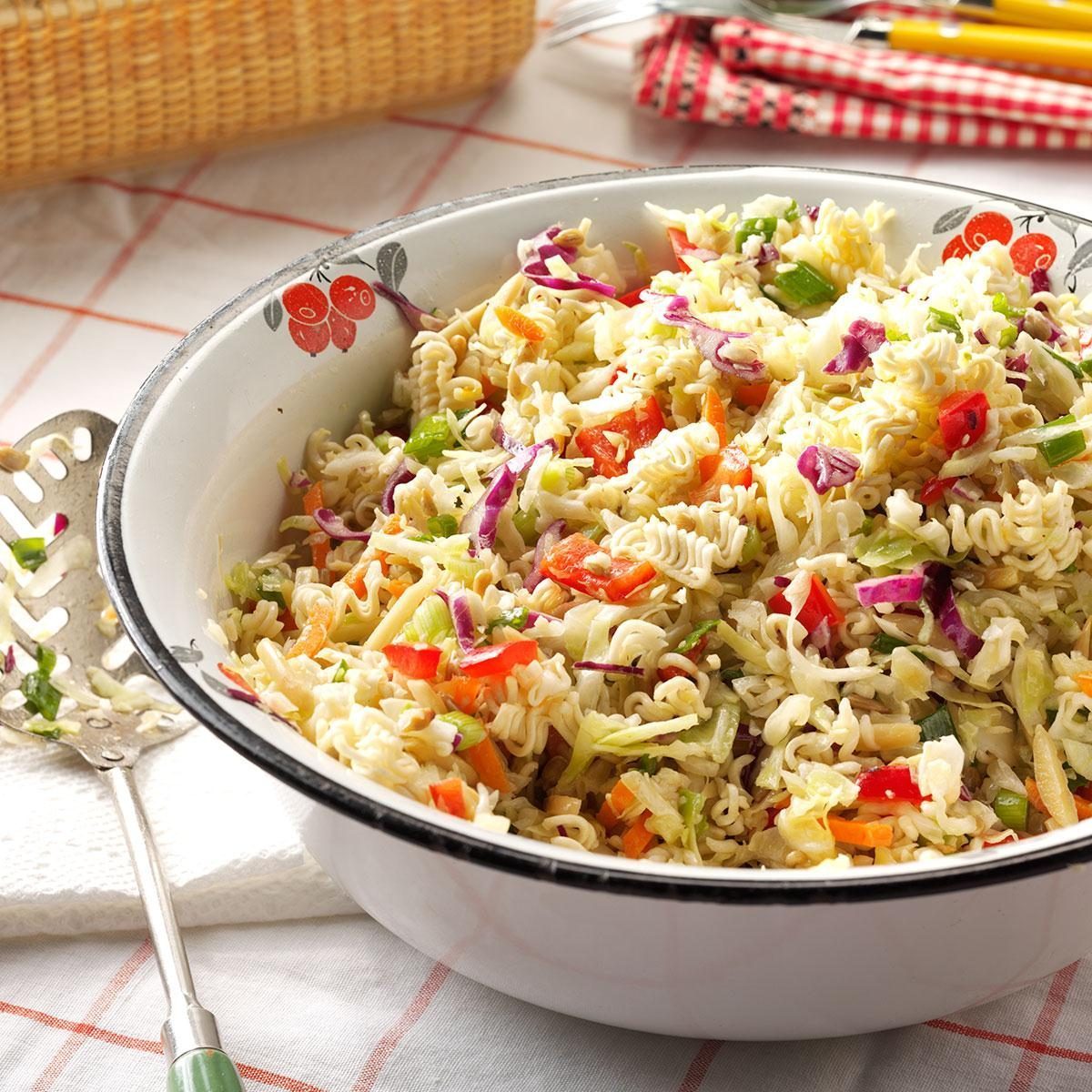 A white bowl with a decorative edge contains a colorful ramen noodle salad, featuring chopped vegetables like red and green bell peppers, purple cabbage, and carrots. A slotted serving spoon rests nearby on a white napkin, with a checkered tablecloth and wicker basket in the background.