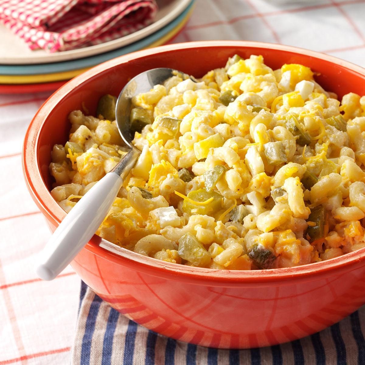 A bowl of creamy macaroni and cheese mixed with diced green peppers is served in a bright orange bowl. A spoon rests in the bowl, ready to serve. The setting includes a checkered tablecloth and stacked plates with a napkin in the background.