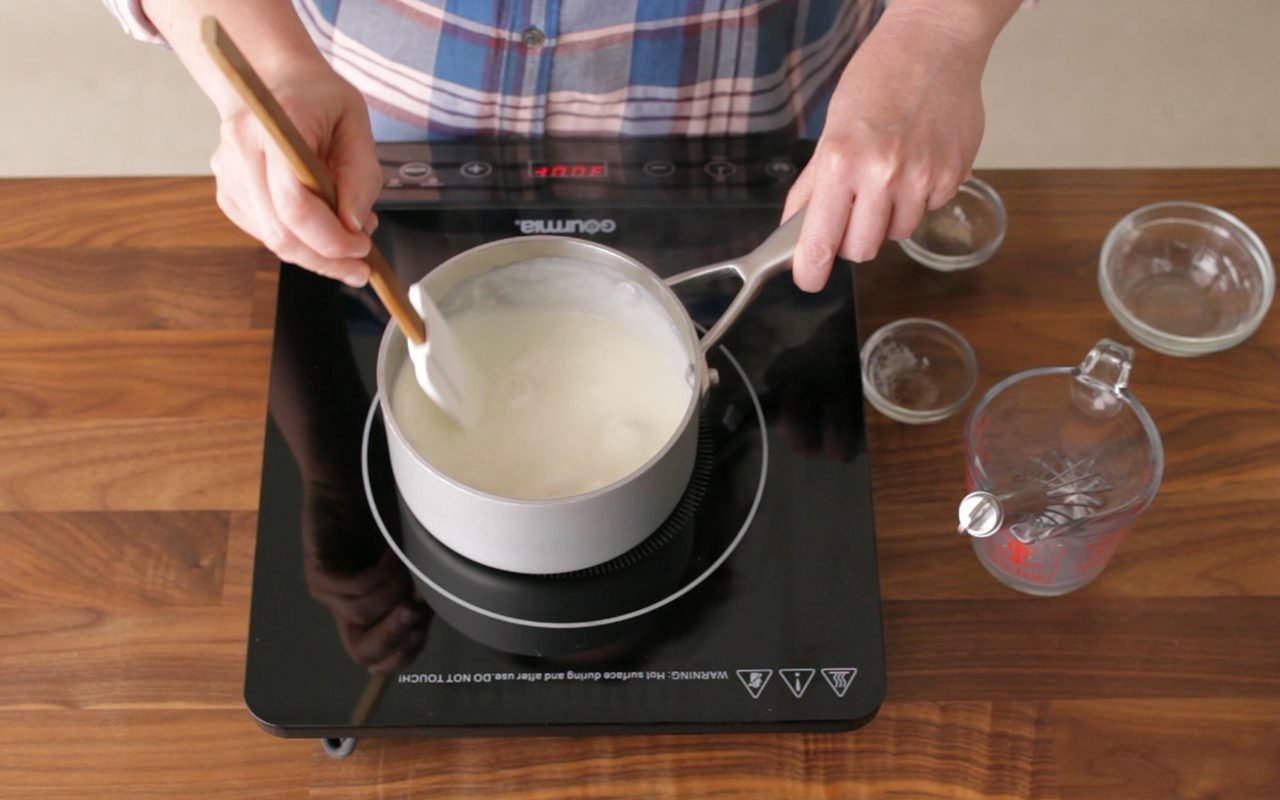 person stirring a white roux with a rubber spatula. roux pan is on a hot plate on a wooden countertop