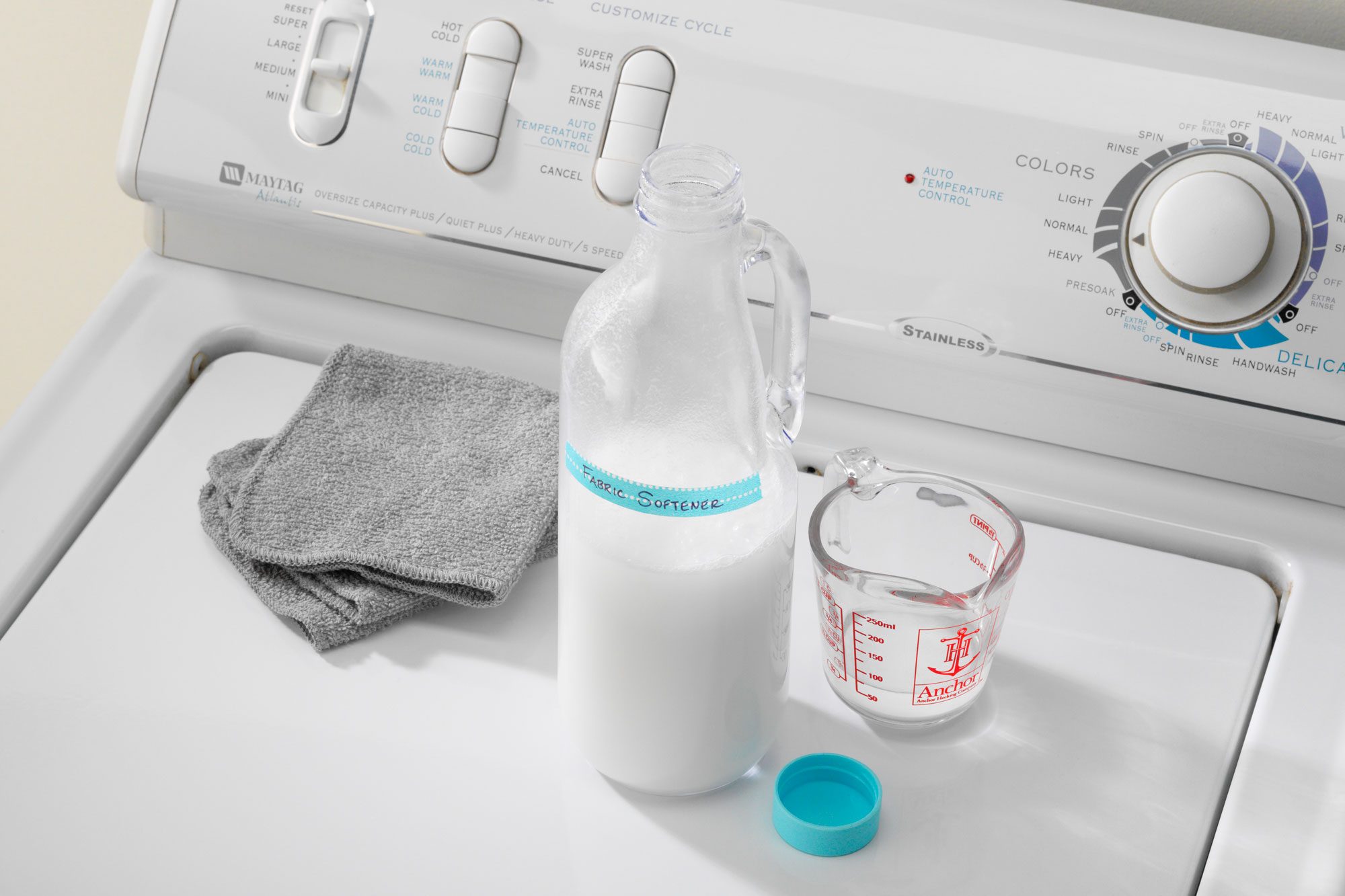 Homemade Cleaners Liquid Fabric Softener in a glass bottle on top of a washing machine next to measuring cup and microfiber cleaning cloth