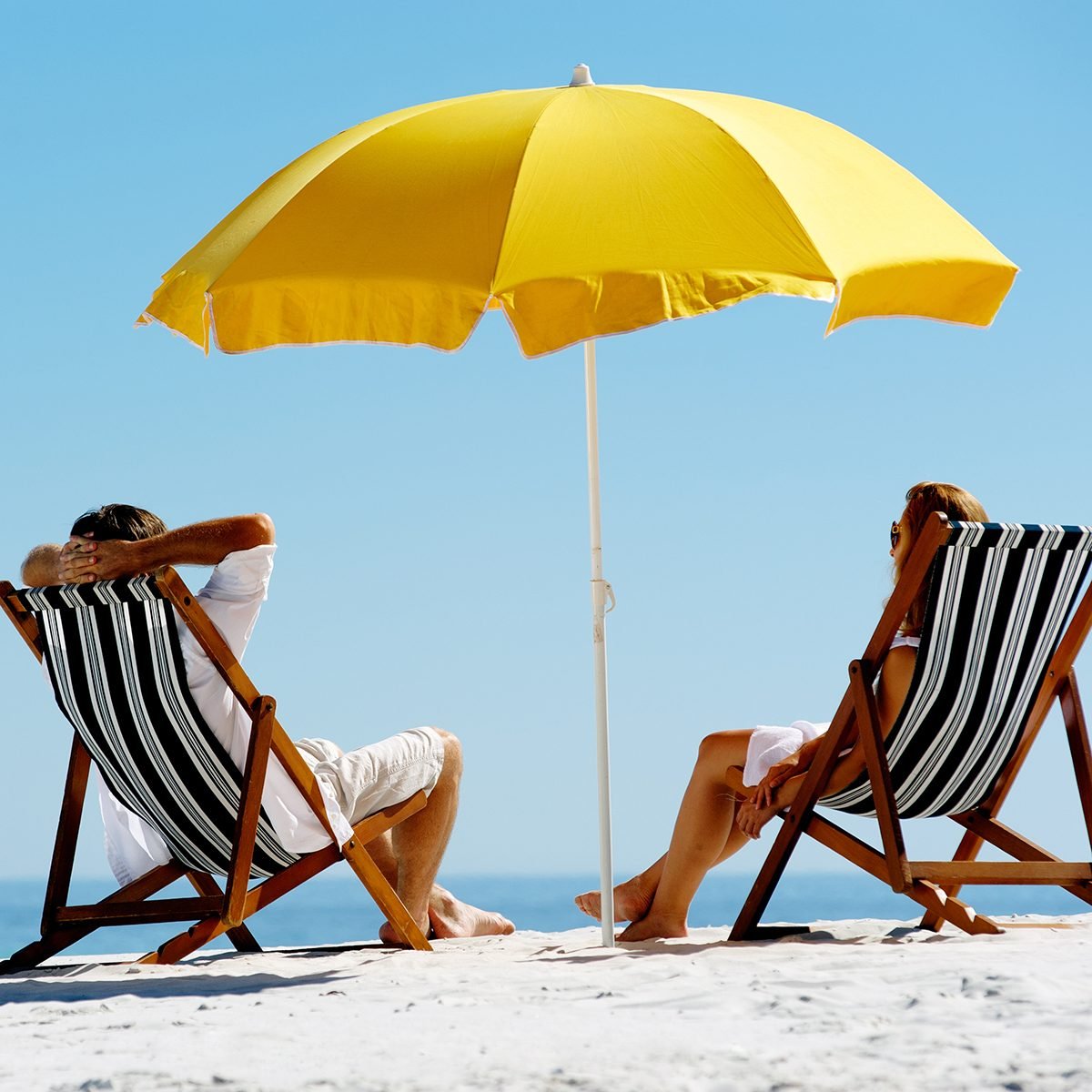 Beach summer couple on island vacation holiday relax in the sun on their deck chairs under a yellow umbrella.