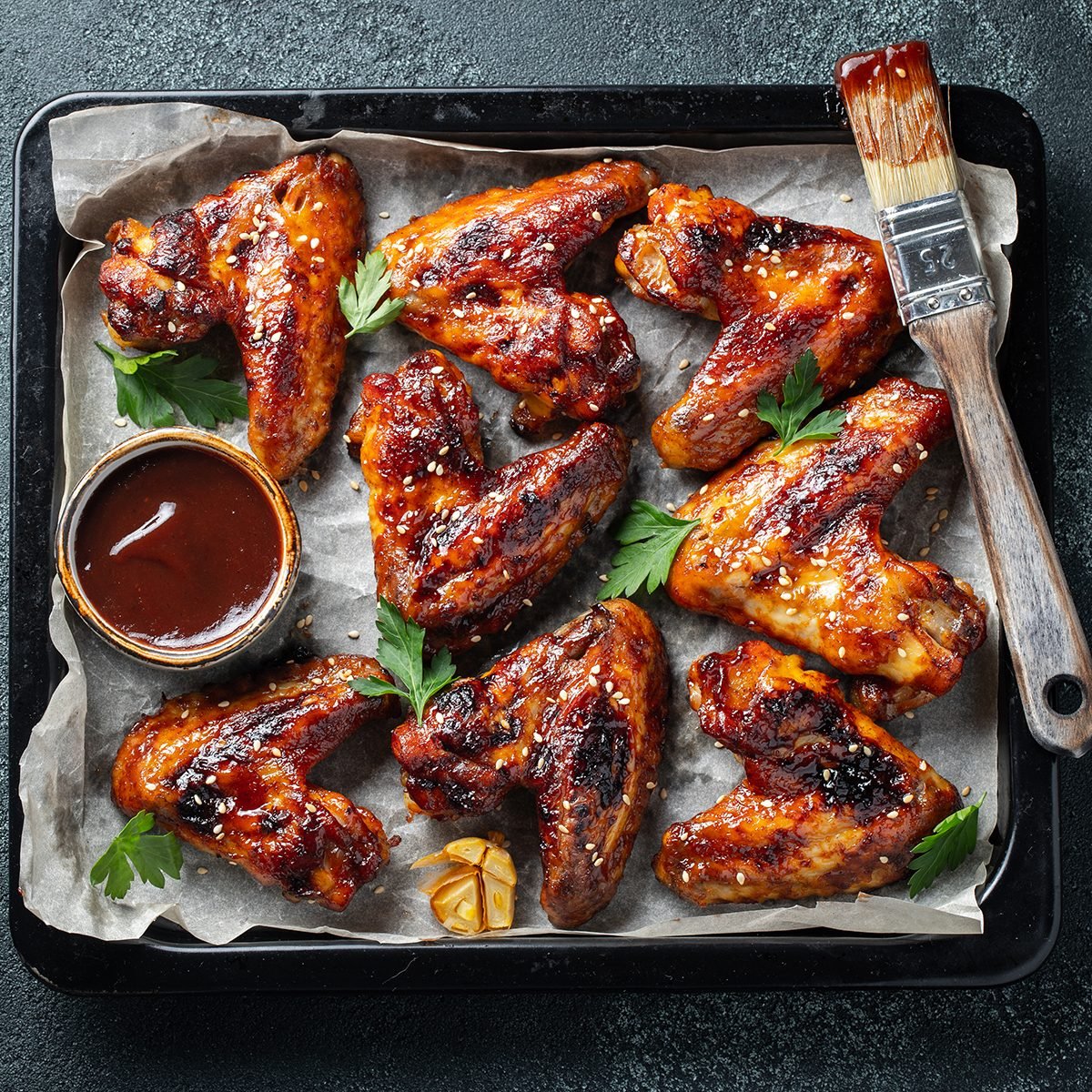 Roasted chicken wings in barbecue sauce with sesame seeds and parsley in a baking tray on a dark table.