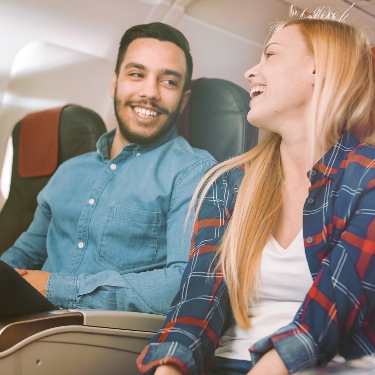 On a Commercial Plane Flight Handsome Hispanic Man Tells Funny Story to His Beautiful Blonde Girlfriend. Both Laugh. They Travel in New Airplane, with Sun Shining Through the Window.; Shutterstock ID 1161853969