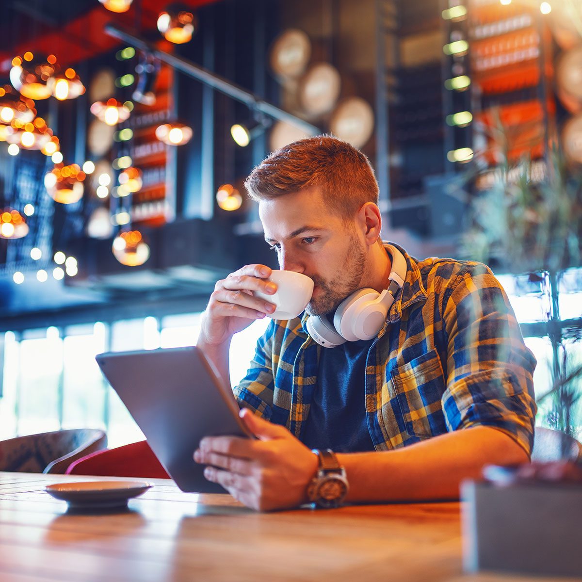 Young handsome Caucasian unshaven man in plaid shirt and with headphones around neck using tablet and drinking espresso while sitting in coffee shop.