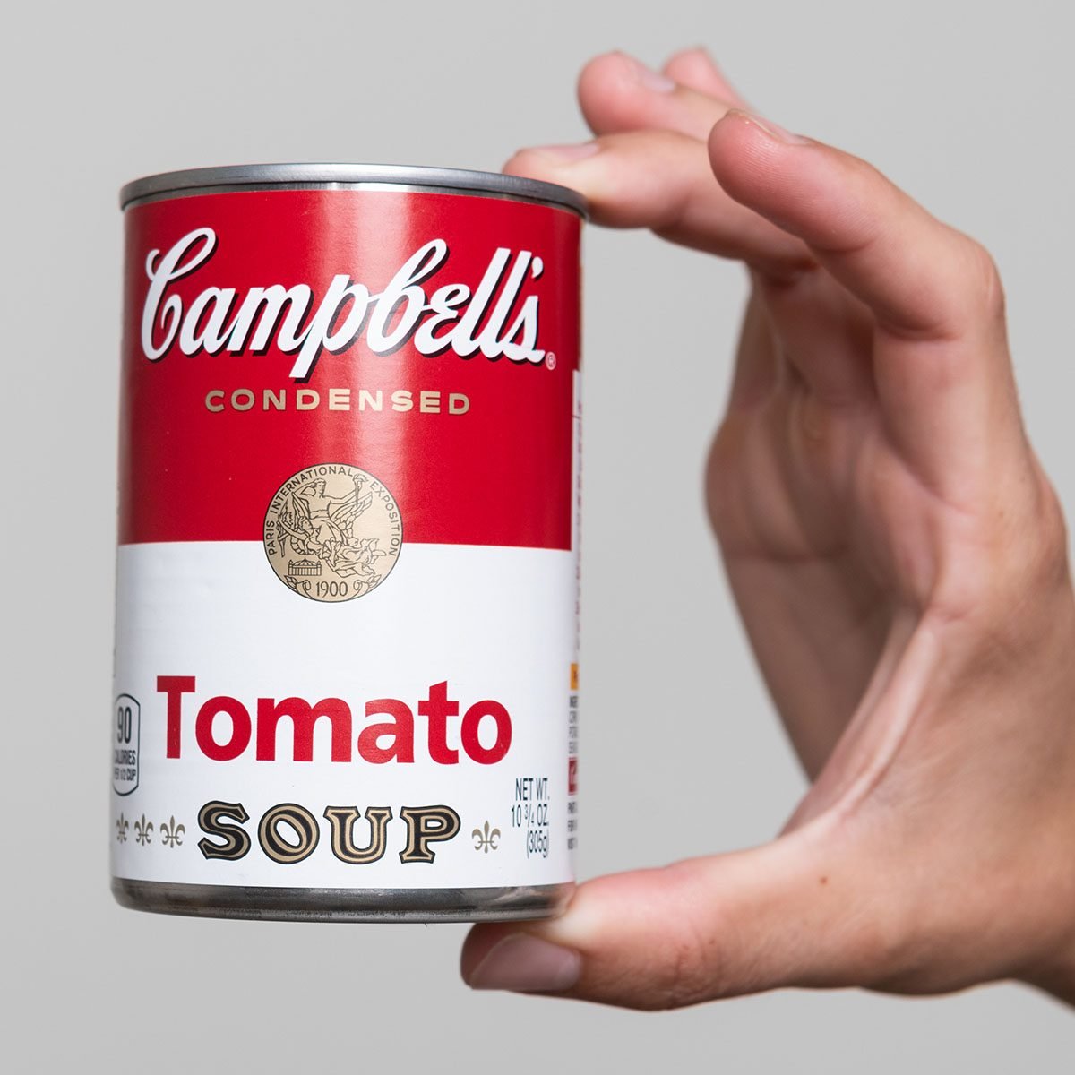 Human hand holding a tin can of Campbell's Soup tomato soup on gray background