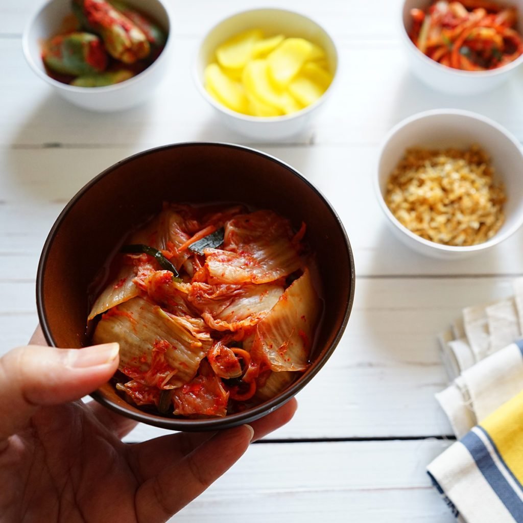 Hand holding bowl of Baechu Kimchi or traditional Napa Cabbage Kimchi over blurred background of colorful variety kimchi and Korean side dishes on white wood planks. Probiotic and fermented foods.; Shutterstock ID 1238289316