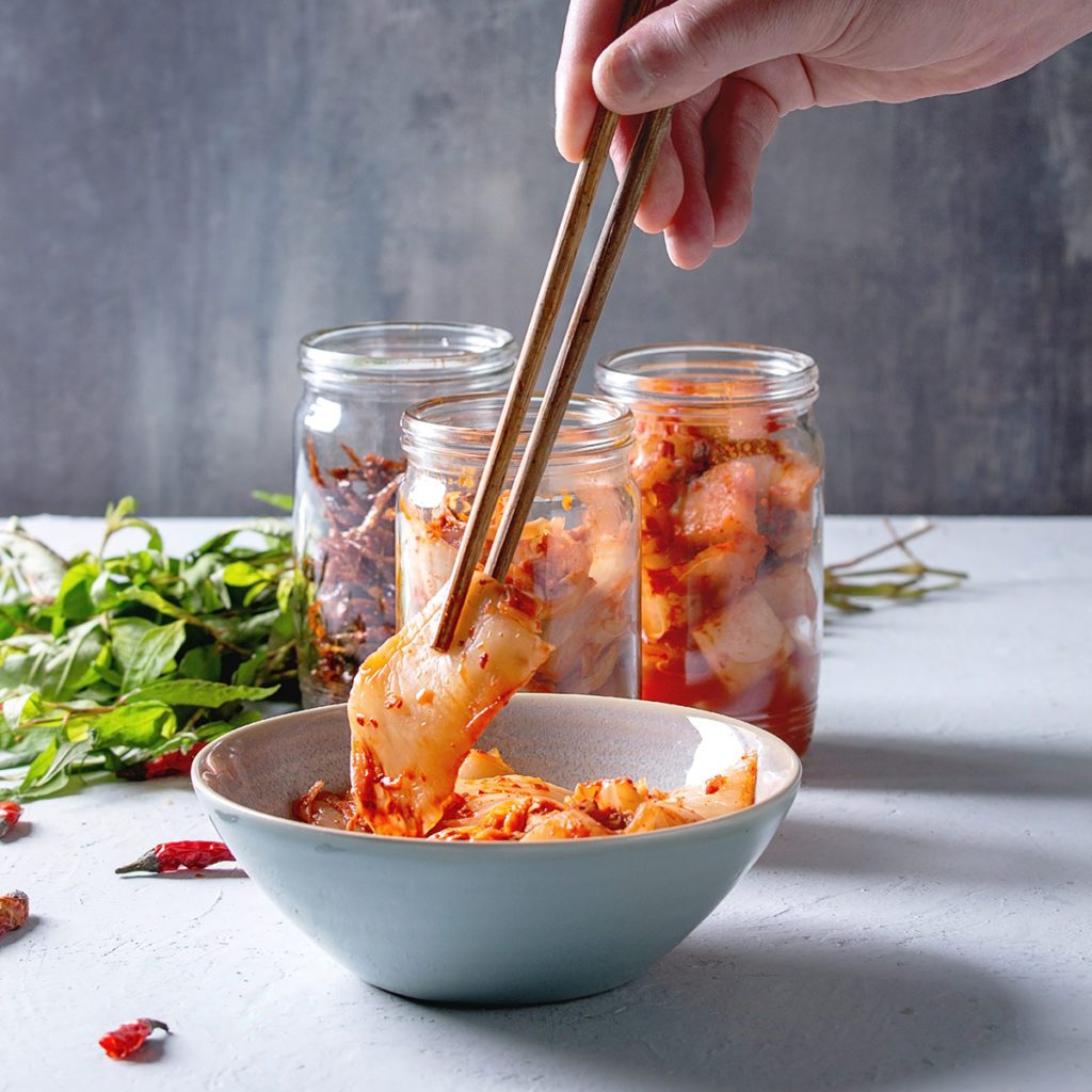 Korean traditional fermented appetizer kimchi cabbage and radish salad, fish snack served in glass jars with Vietnamese oregano and chili peppers over grey blue table. Chopsticks in mans hands.; Shutterstock ID 1345435652; Job (TFH, TOH, RD, BNB, CWM, CM): Taste of Home