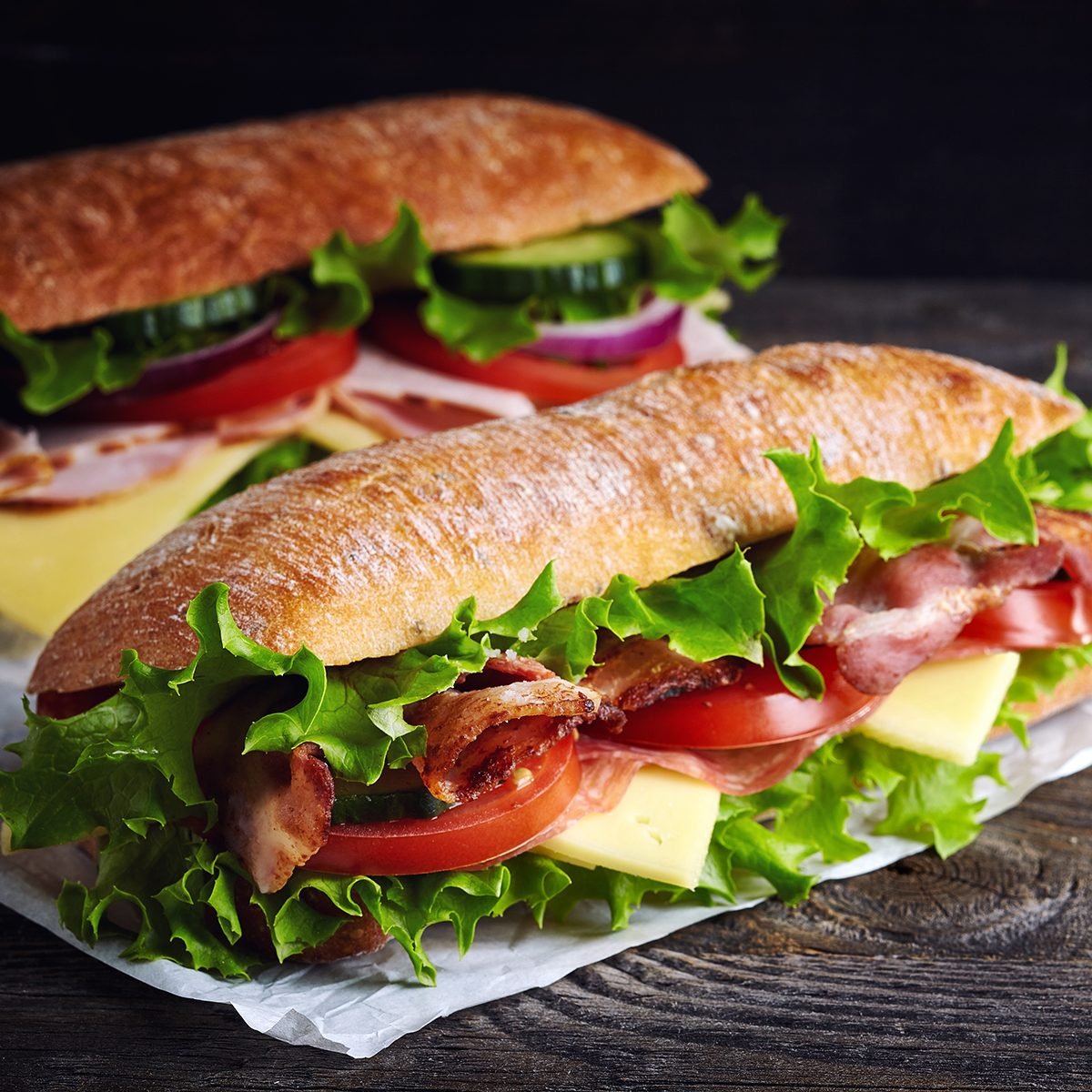 Two fresh submarine sandwiches with ham, cheese, bacon, tomatoes, lettuce, cucumbers and onions on dark wooden background