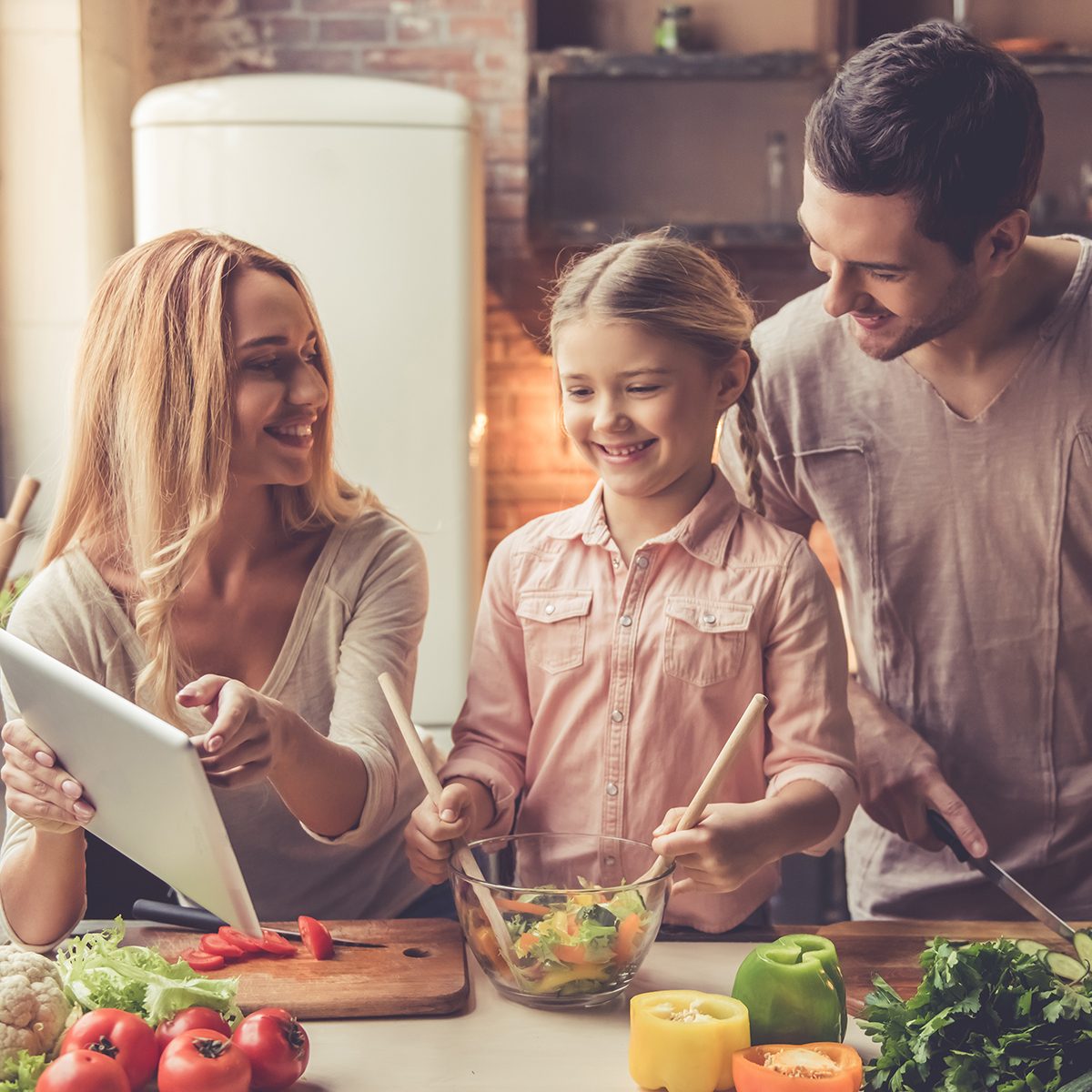 Cute little girl and her beautiful parents are using digital tablet and smiling while cooking in kitchen at home