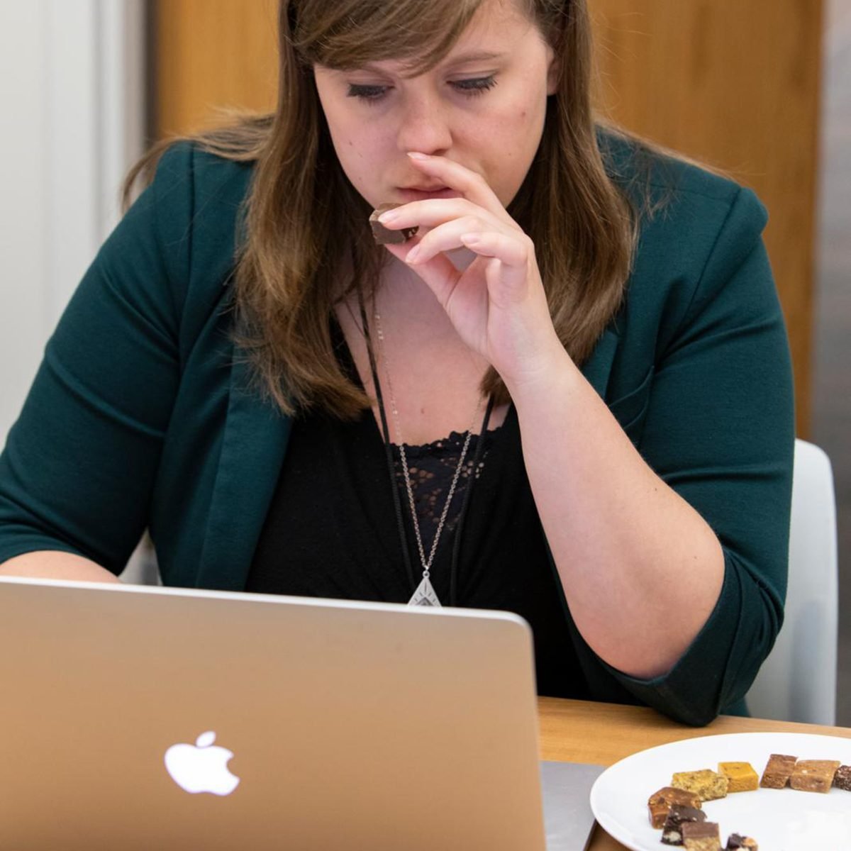 Woman eats small piece of protein bar while looking at laptop