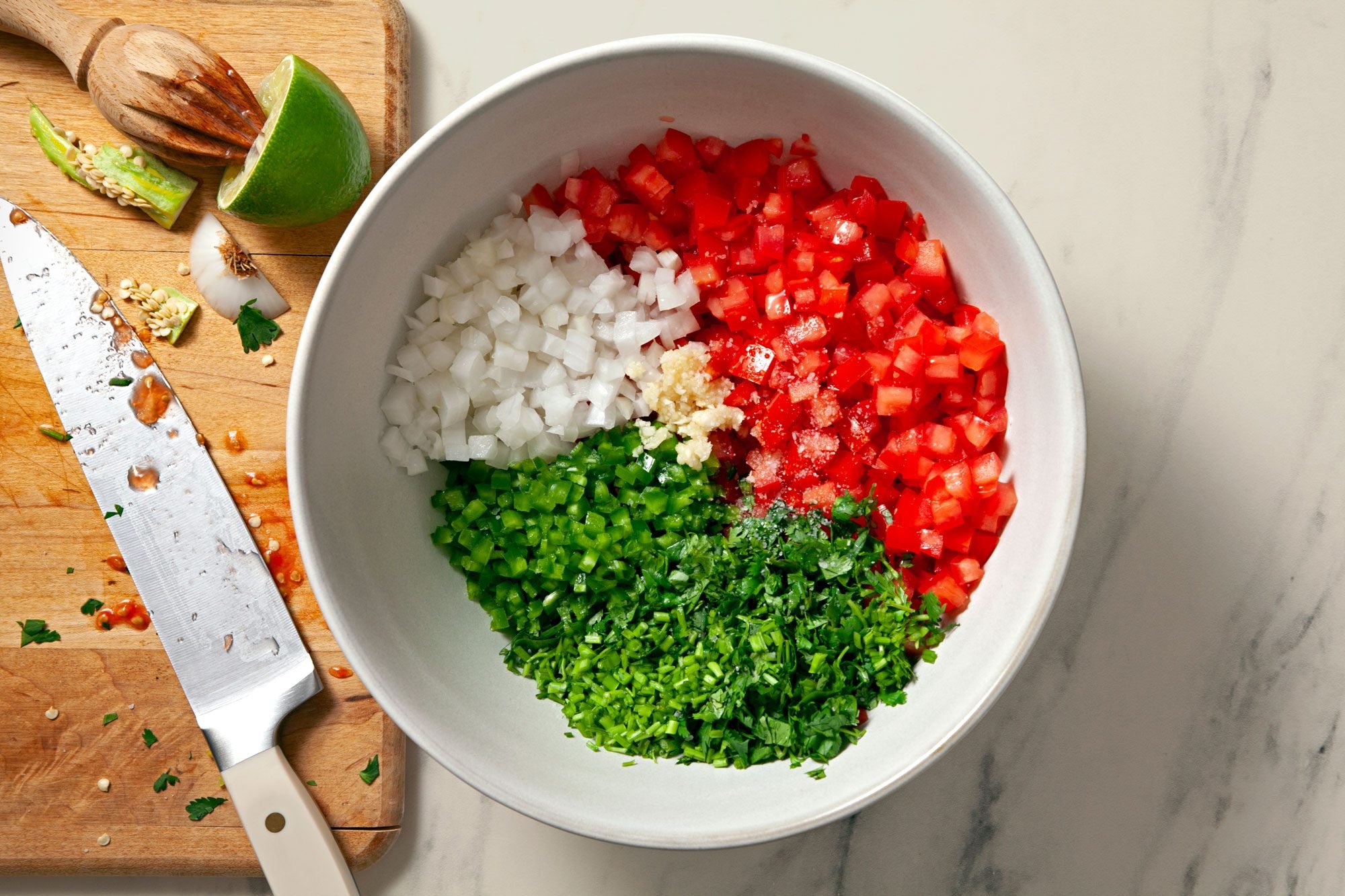 Overhead shot of a large bowl; chopped tomatoes; onion; fresh cilantro; jalapeno peppers; seeded and lime juice; 1 tablespoon cilantro stems; finely chopped 1 garlic clove; knife; wooden chopping board; marble background;