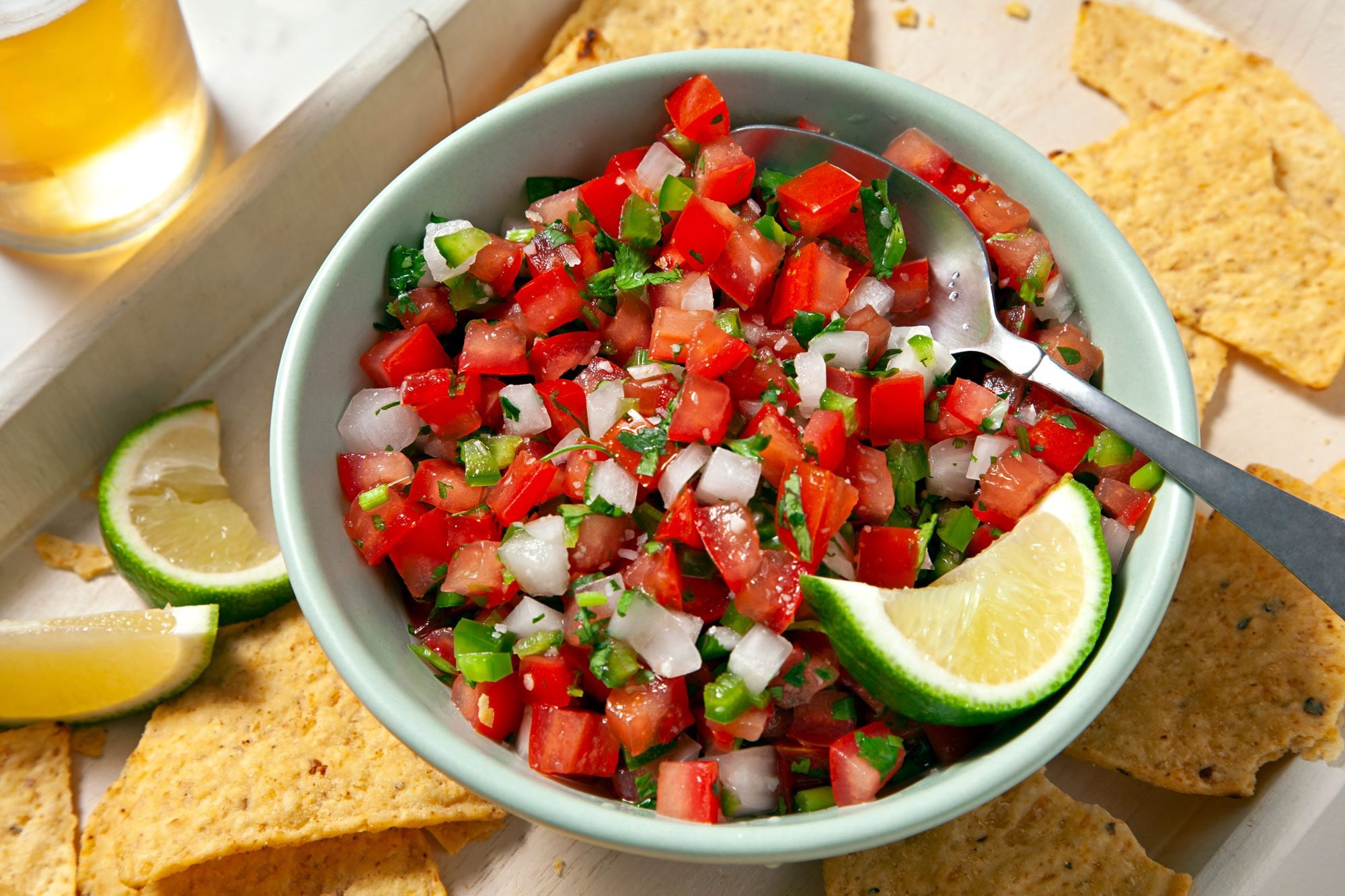Close shot of Pico De Gallo; in a bowl; top with lemon wedge; served with nachos; large platter; a glass of drink; spoon; marble background;