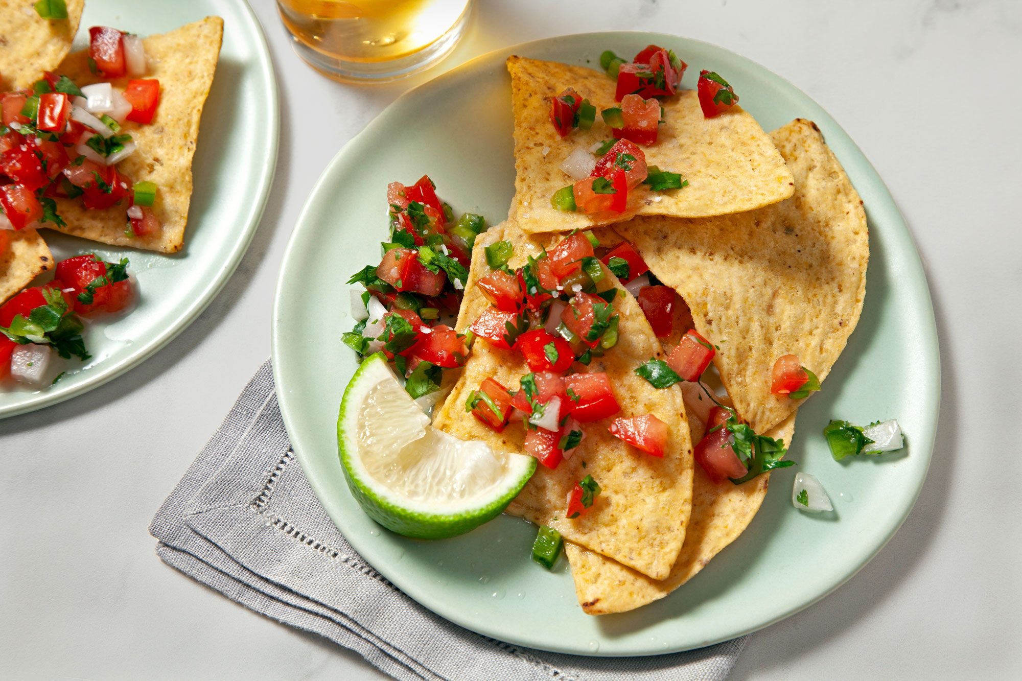 Overhead shot of Pico De Gallo; served on two white plates; with nachos and top with lemon wedge; napkin; a glass of drink; marble background;