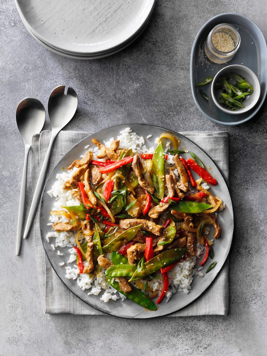 Ginger Pork Stir-Fry; overhead camera angle; mottled grey Russian surface; Grey linen cloth; grey folded napkin; salad serving forks; Grey plate stack; green onions; sesame seeds; snow peas; sweet red pepper; white rice