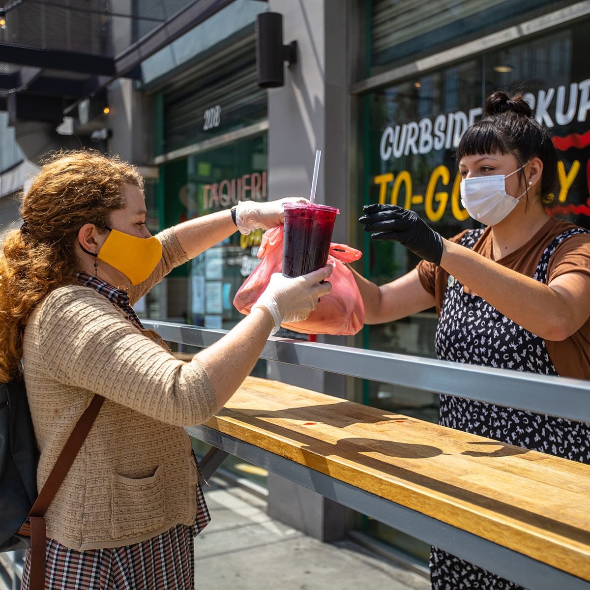 A Mexican restaurant adapts to the Covid-19 lockdown. The owner hands an order to a customer outside the restaurant; they are both wearing gloves and masks.