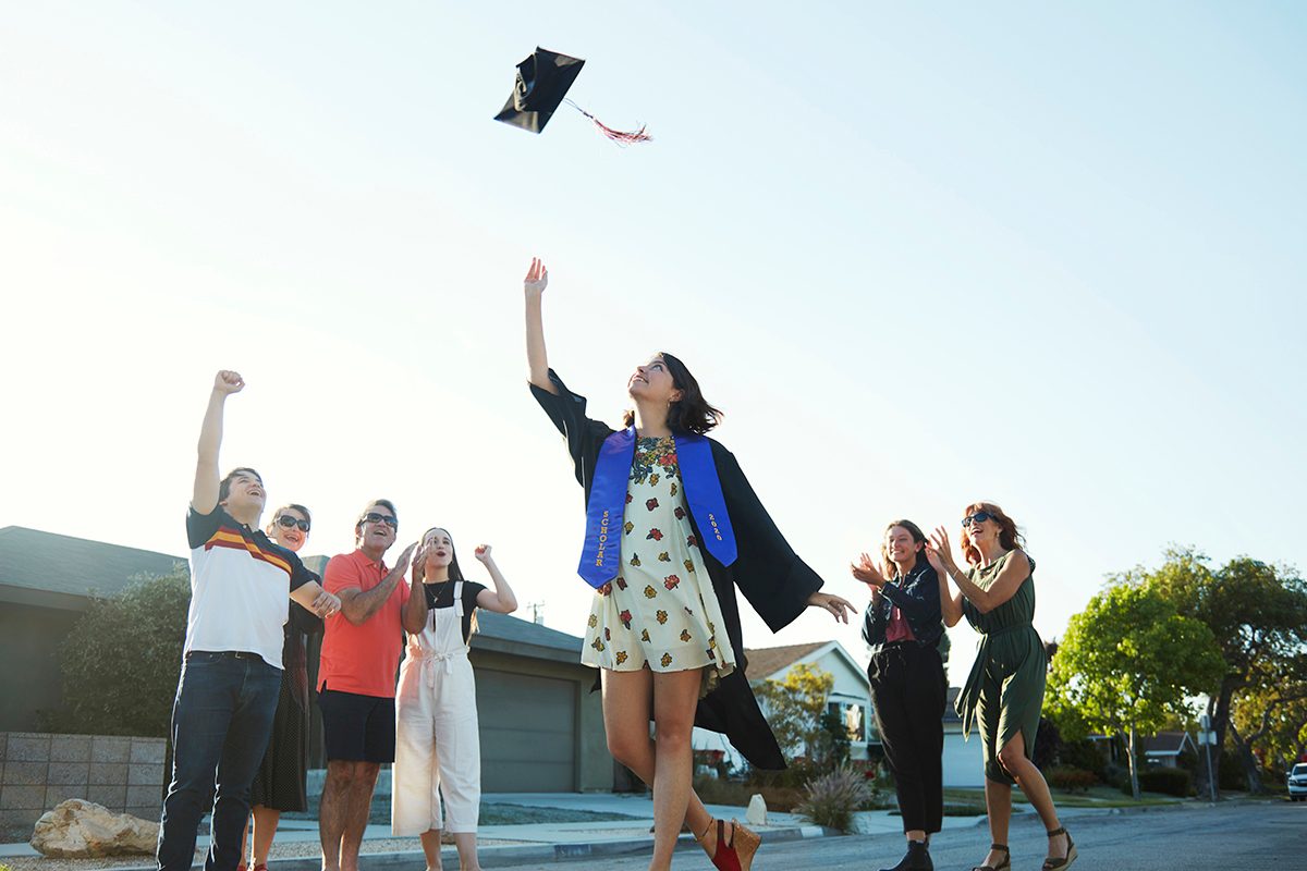 A 22 year old caucasian young woman dressed in a graduation gown throws her hat into the air surrounded by her friends and family in front of her home while celebrating her graduation from college