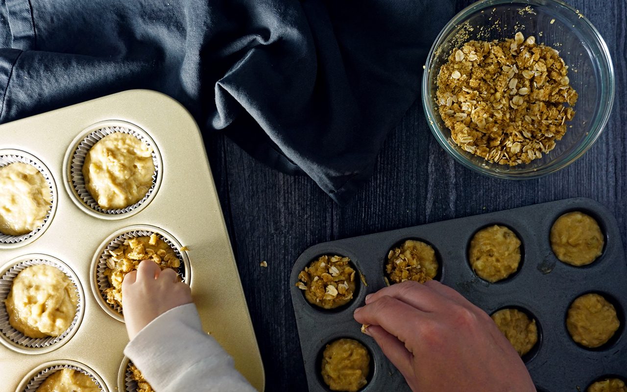 sprinkling streusel topping with a toddler over healthy muffin batter in standard and mini muffin tins