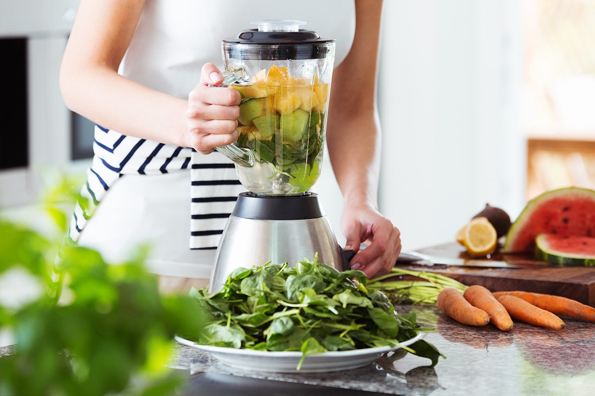 Vegetarian preparing vegan smoothie with rucola, citrus, cucumber in kitchen with carrots on countertop