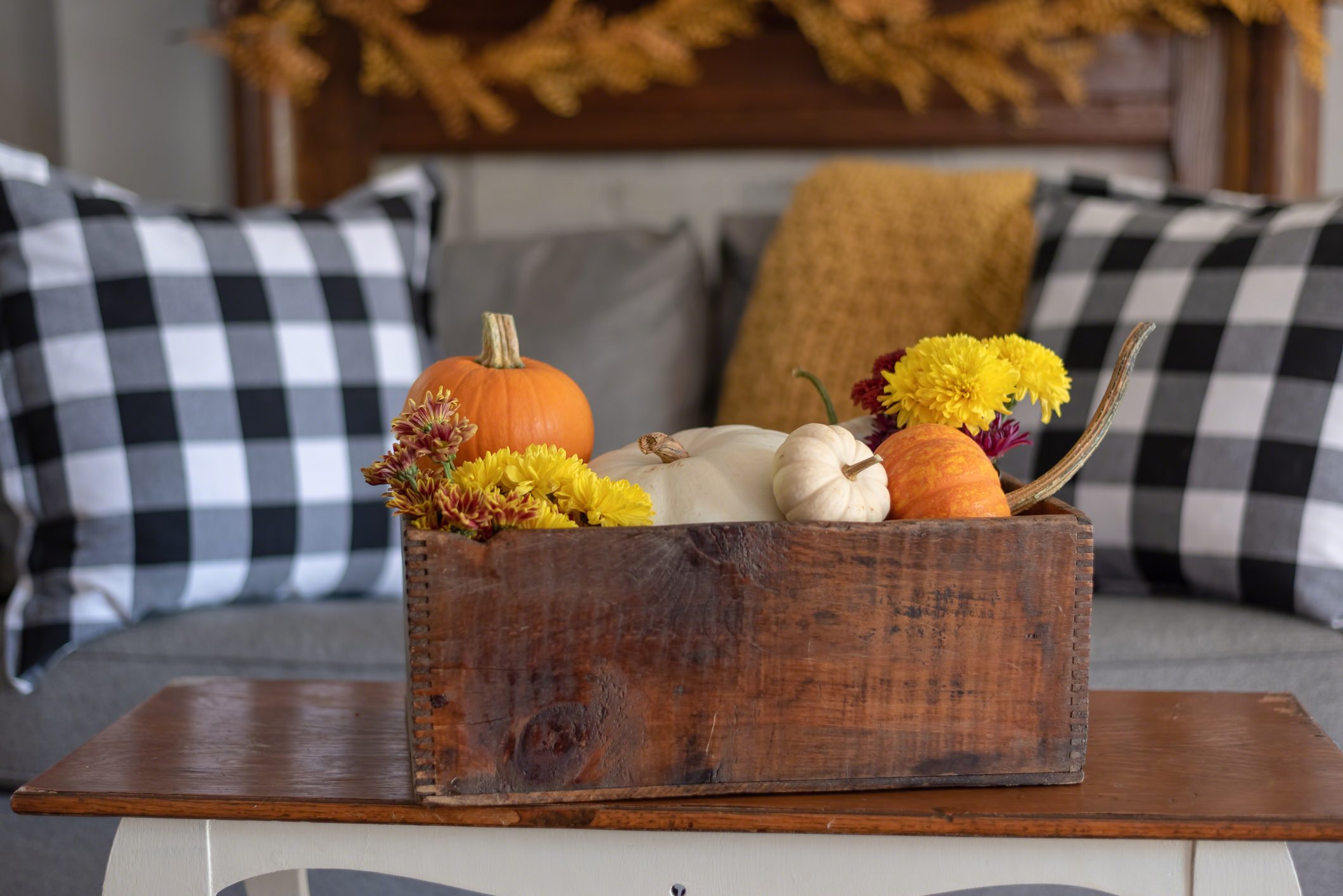 Vintage wooden crate filled with pumpkins and autumn flowers on a coffee table; couch with gingham pillows and a yellow throw blanket in the background