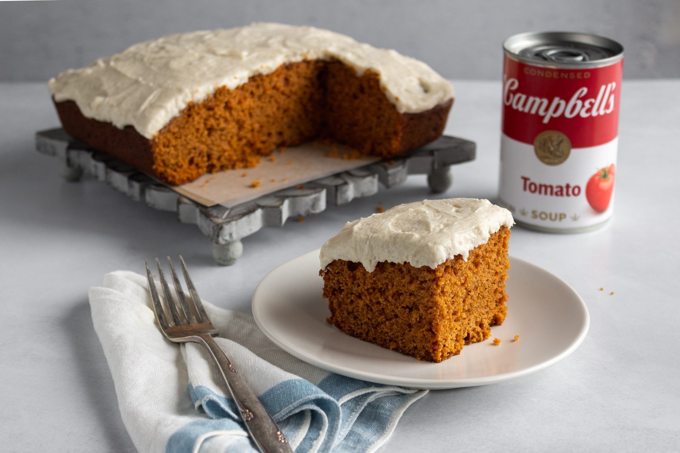 Tomato Soup Cake slice on a white place with full cake in the background next to a can of campbells tomato soup