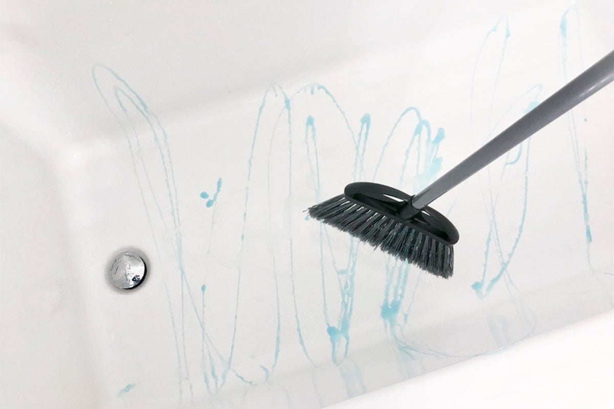 dark gray broom being used to scrub dish soap around to clean a bathtub