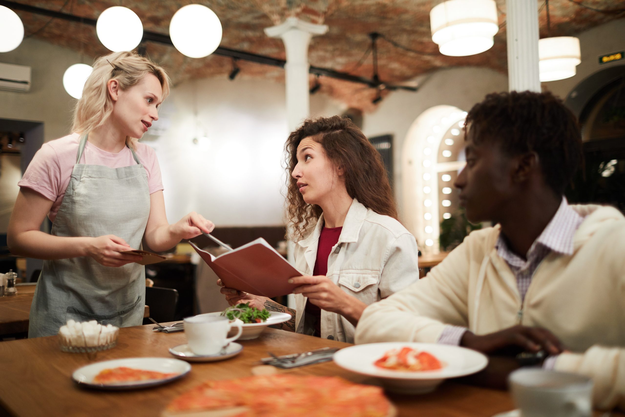 Disgruntled Curly Haired Young Female Restaurant Guest Sitting At Table With Boyfriend And Reading Menu While Talking To Waitress Giving Advice To Her In Modern Loft Cafe