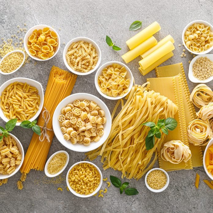 Various kinds of uncooked pasta and noodles, some in white bowls, on stone background, top view