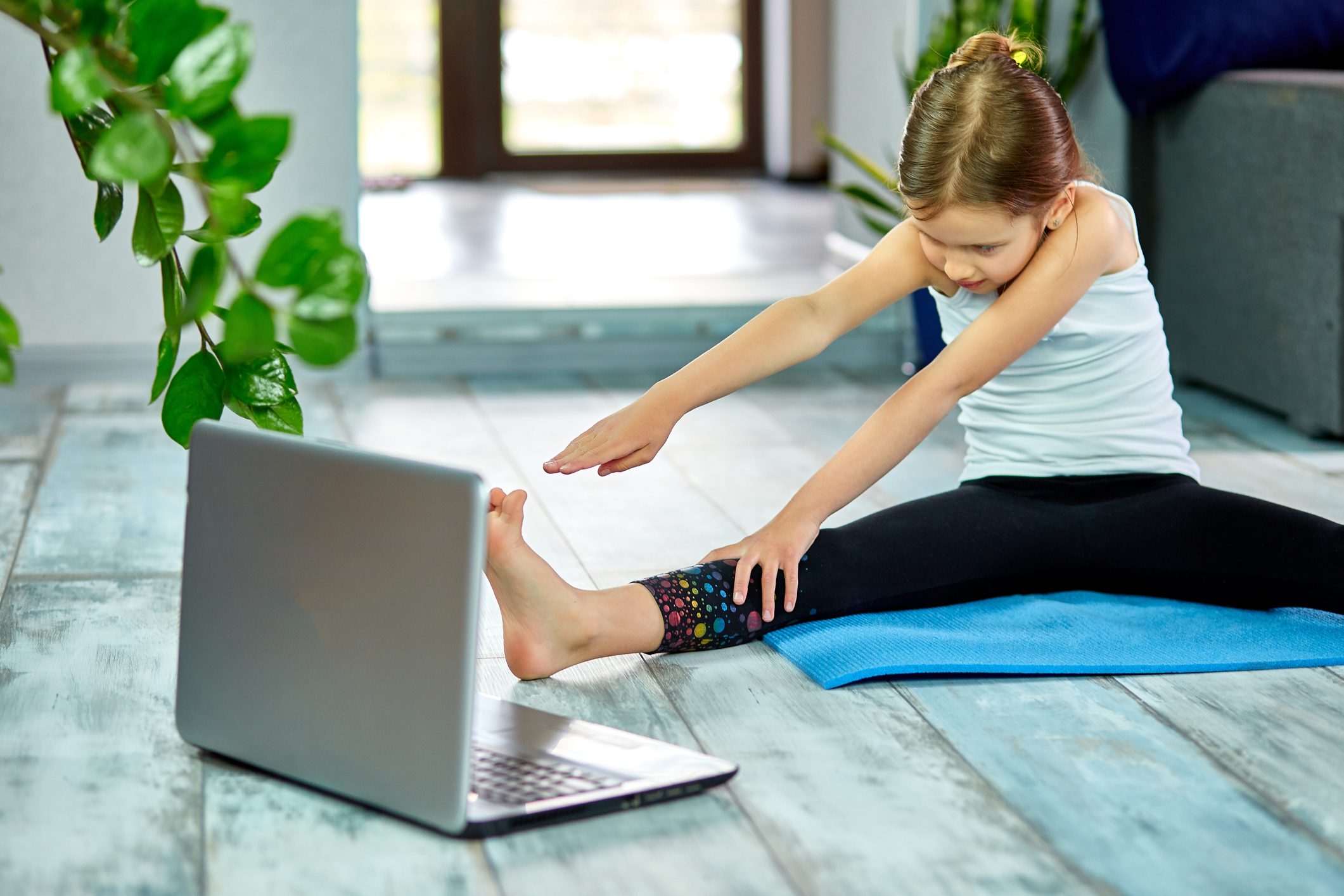 Little girl make workout on blue mat at home by laptop online.