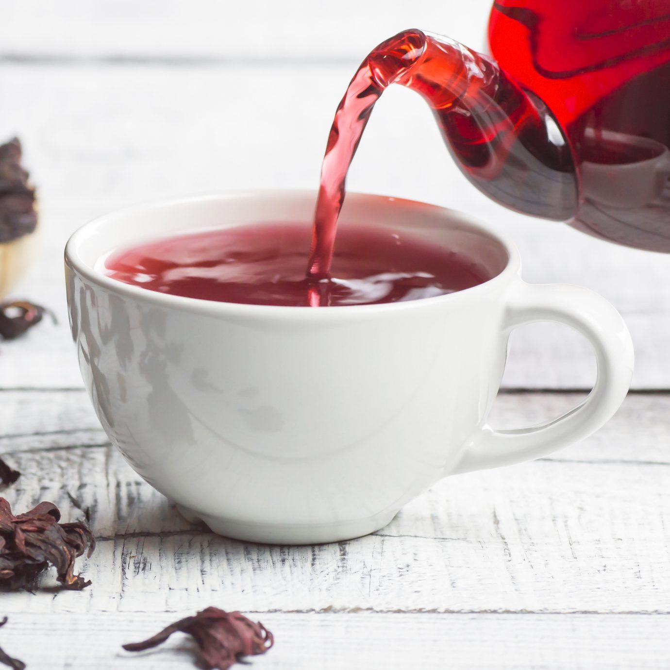 White cup of healthy hibiscus tea pouring from the teapot with dried hibiscus flowers on white wooden background, winter hot drink concept for cold and flu