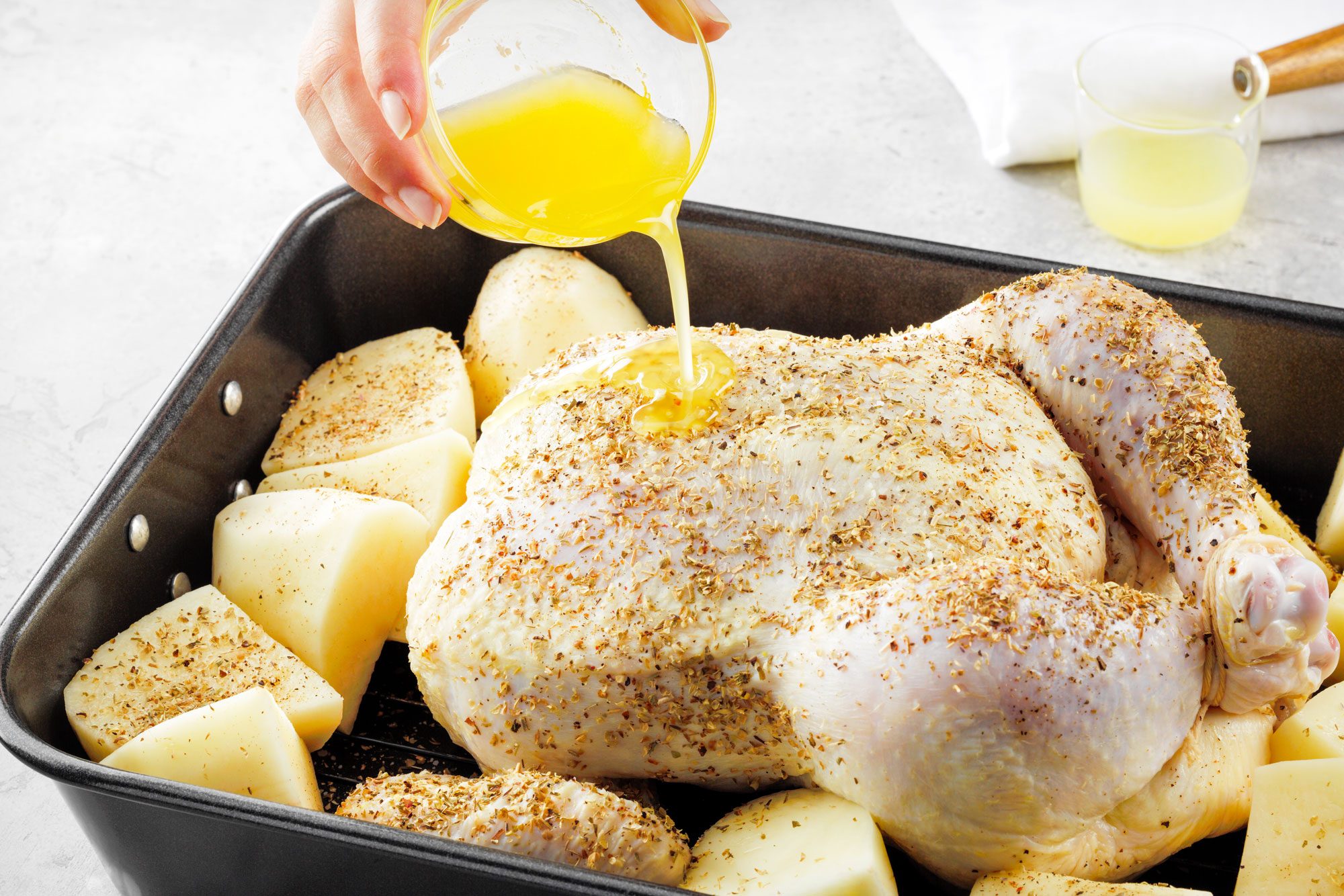 pouring melted butter over chicken and potatoes placed in a roasting pan on marble surface
