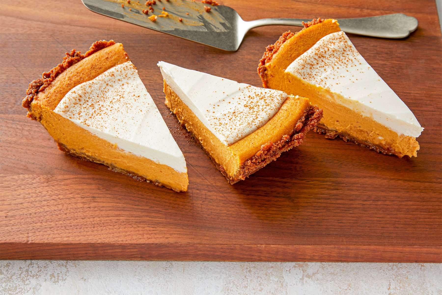 Three slices of pumpkin pie topped with whipped cream and a sprinkle of cinnamon are displayed on a wooden surface. A serving spatula with some remnants of the pie is placed beside them.