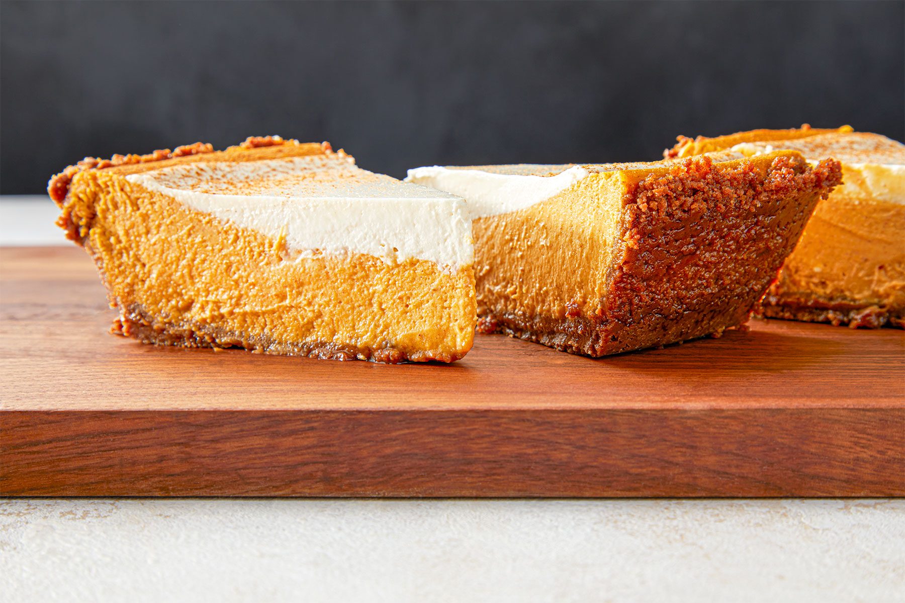 Two slices of pumpkin cheesecake with a graham cracker crust and a layer of whipped topping are placed on a wooden surface. The background is dark, providing contrast to the dessert.