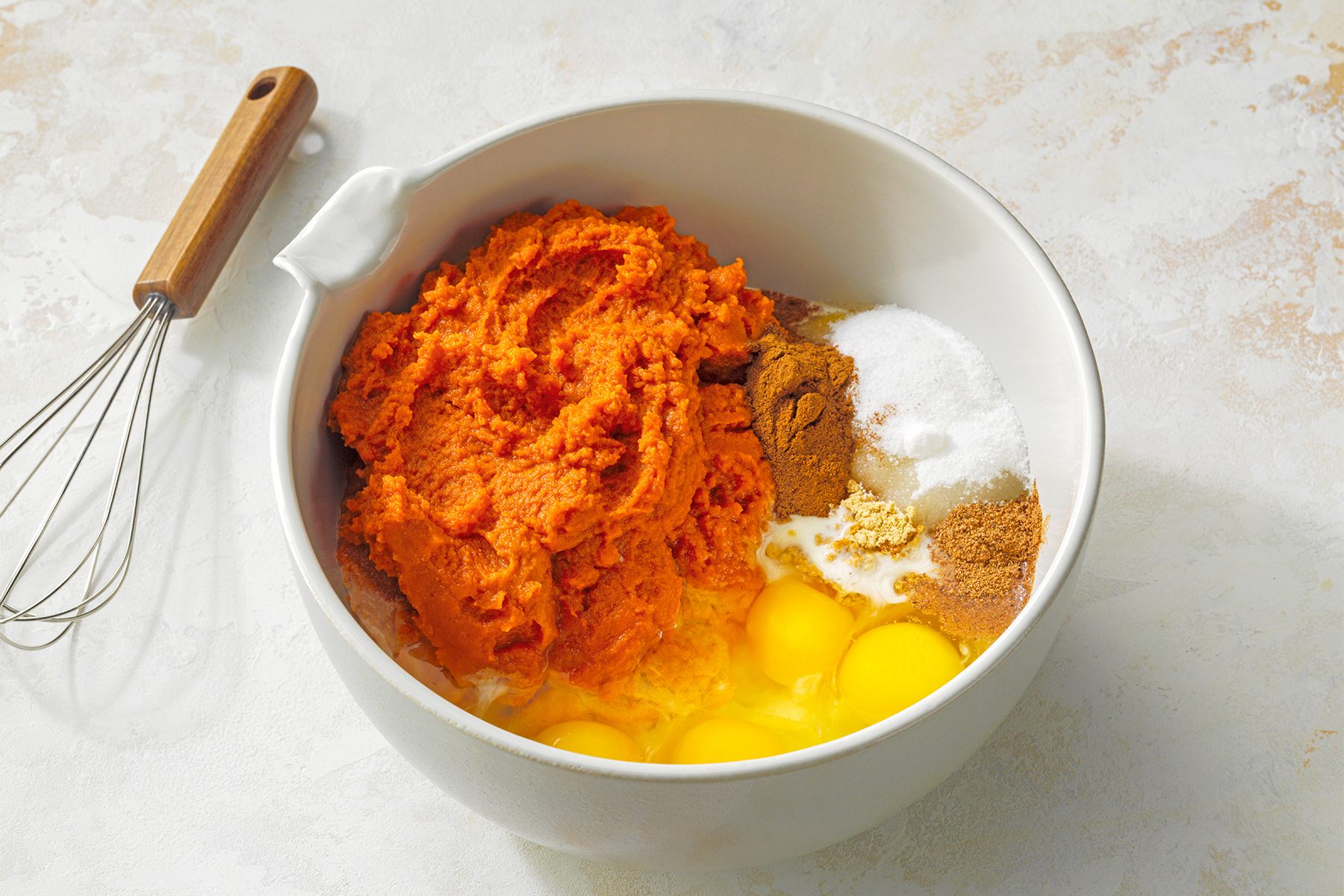 A large white mixing bowl contains a combination of orange pumpkin puree, four cracked eggs, granulated sugar, ground cinnamon, nutmeg, and a brown spice