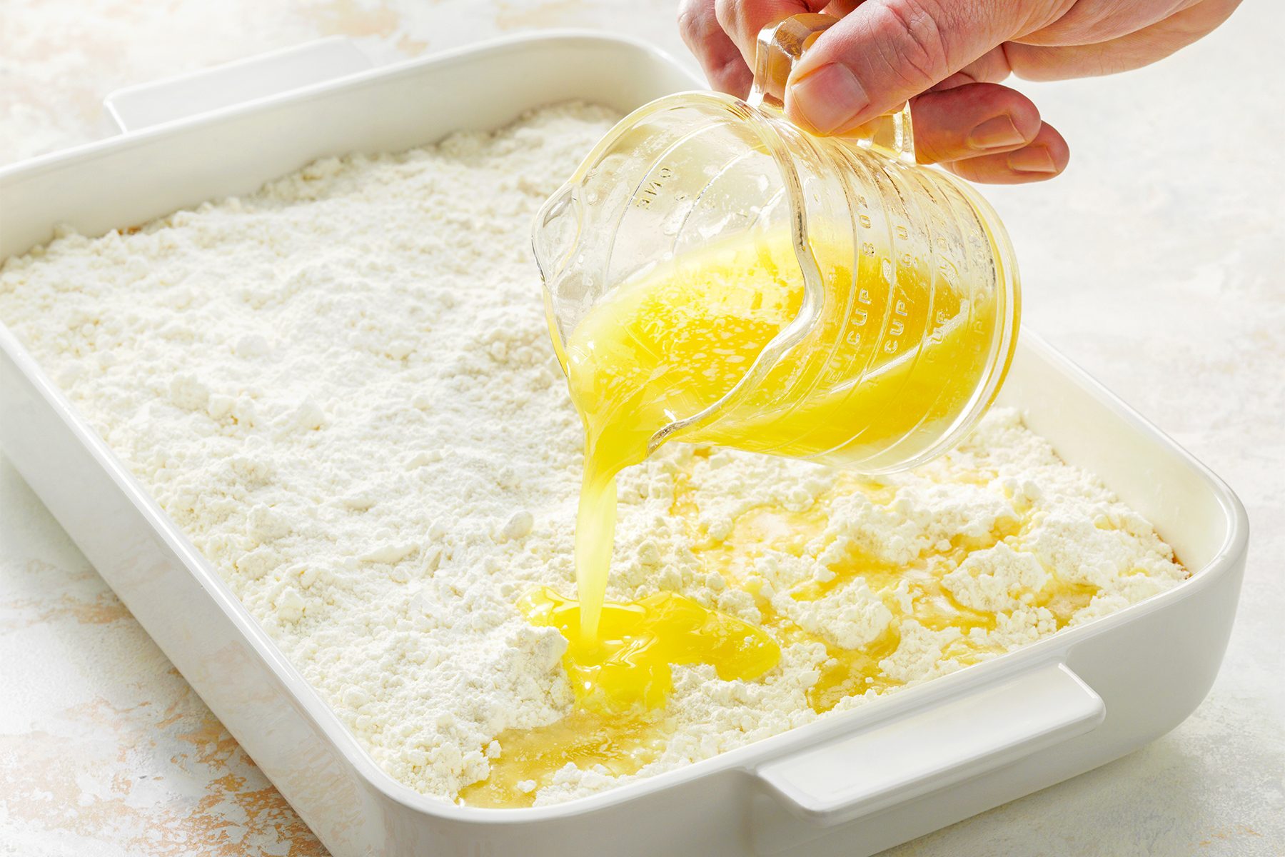 A hand is pouring melted butter from a measuring cup into a rectangular white baking dish filled with flour