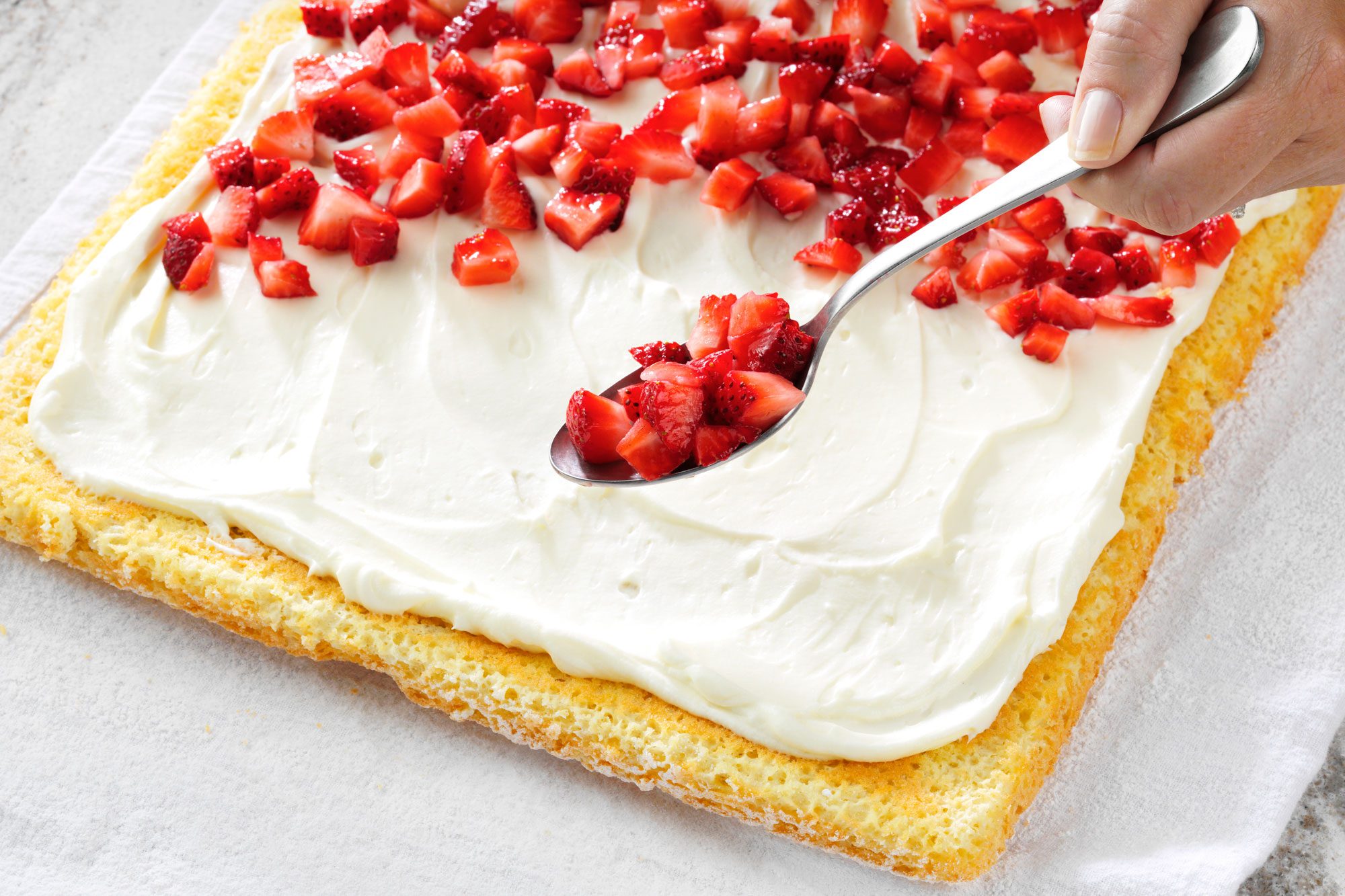 tilted shot; white textured background; Sprinkling strawberries over whipped cream spread on cake with silver spoon;