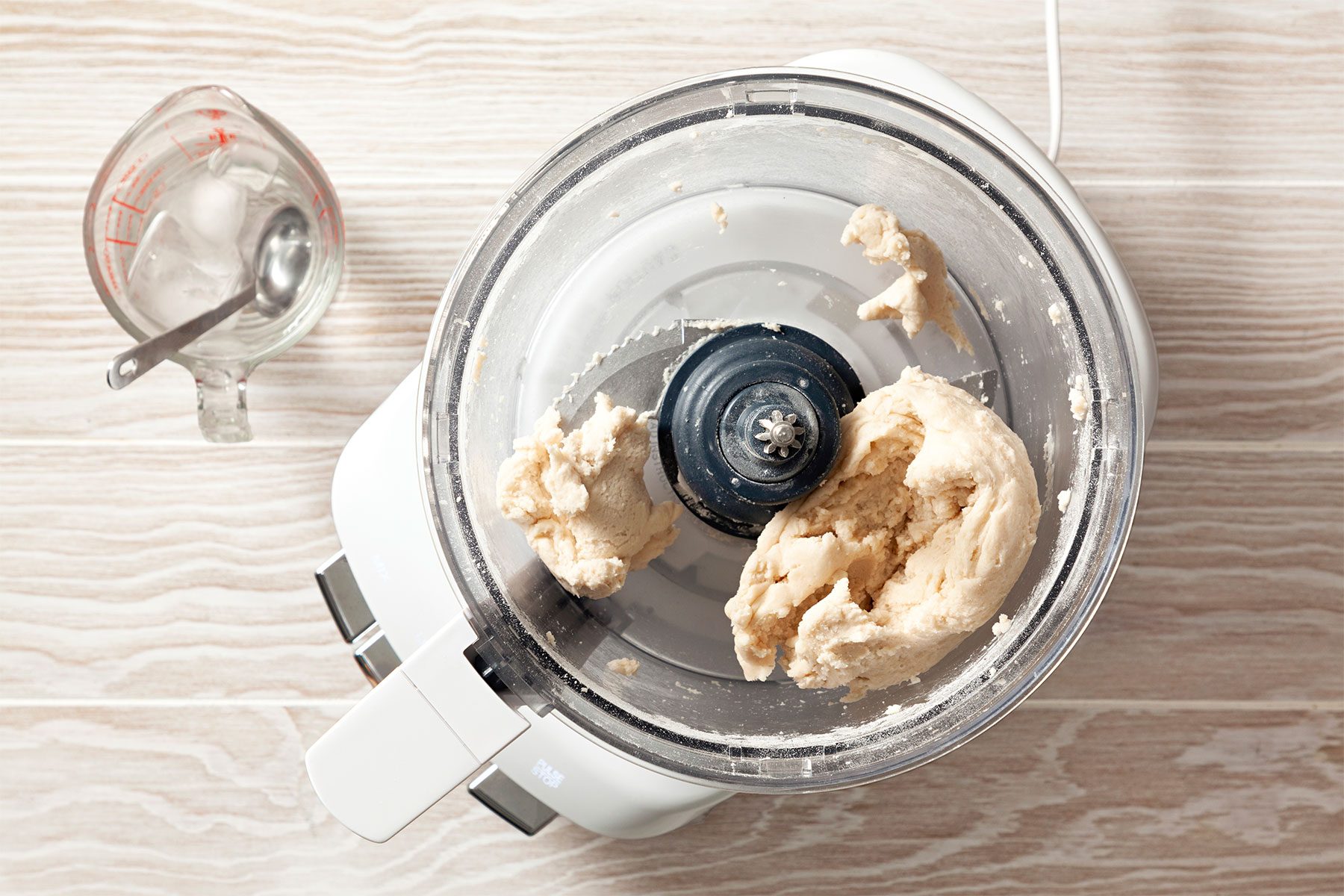 overhead shot; wooden background; In a food processor, mixing flour to form dough; ice water in a beaker with small spoon;