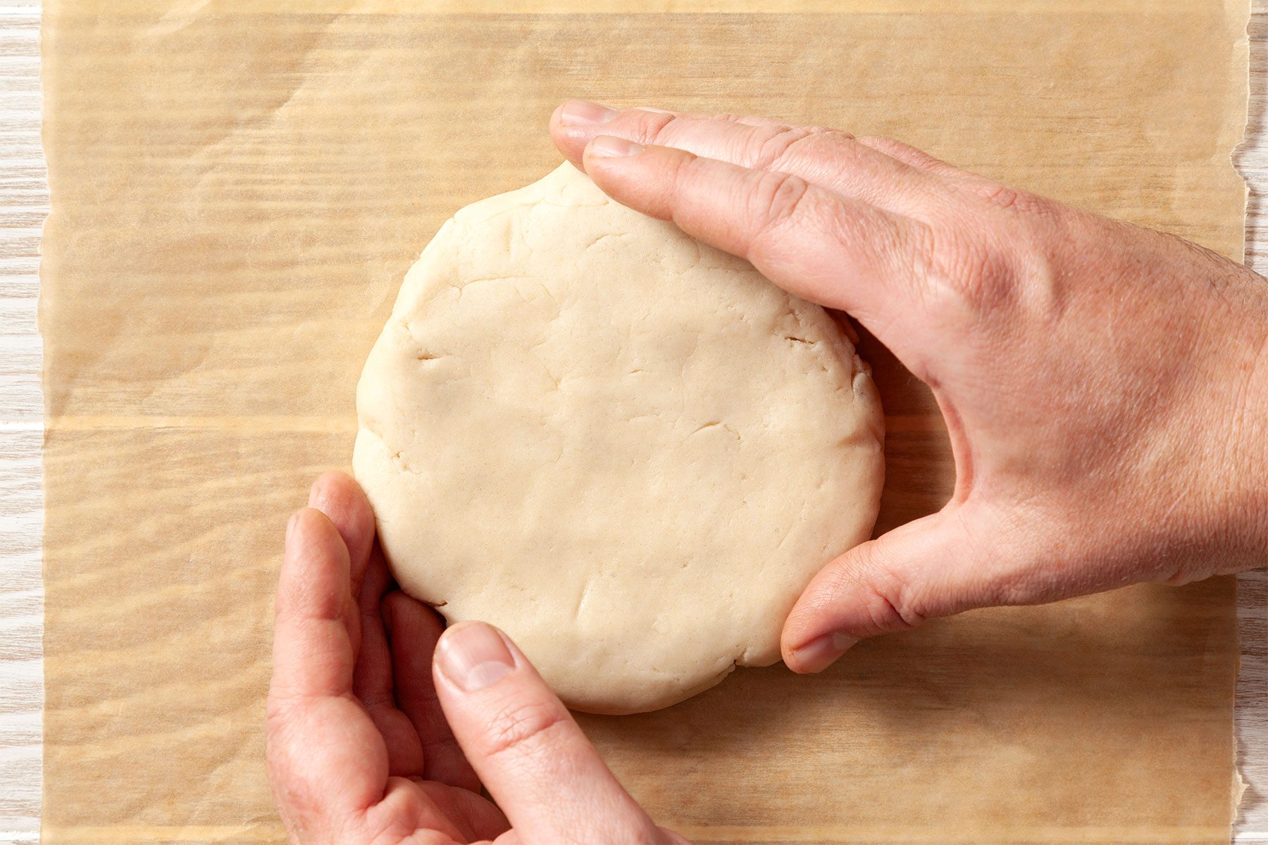 overhead shot; wooden background; Shaping dough into a disk with hands over wax paper;
