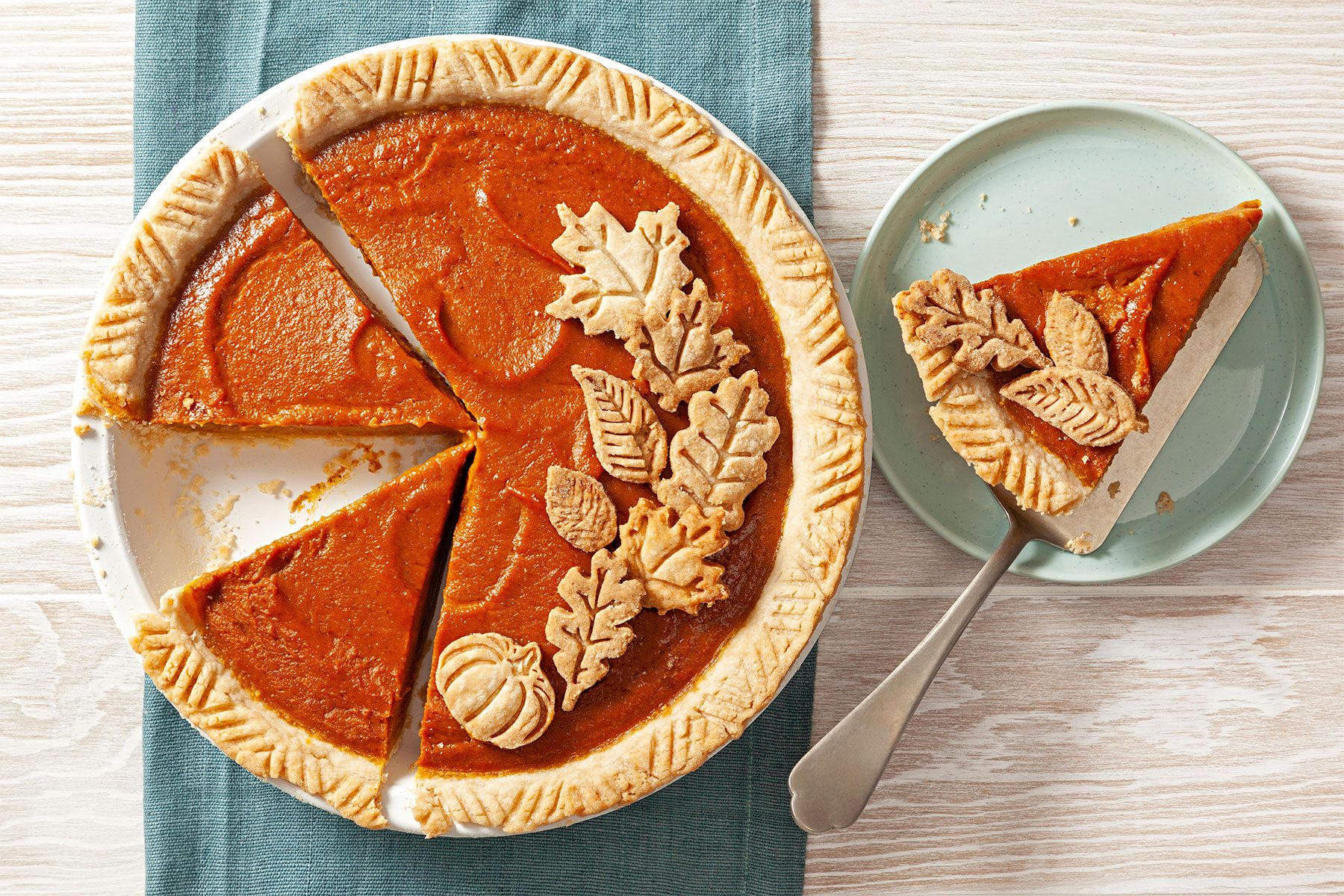 overhead shot; horizontal shot; wooden background; Pumpkin Pie in pie plate over green kitchen towel; garnishned with leaves cutout; pie piece served on small green plate with small spoon;