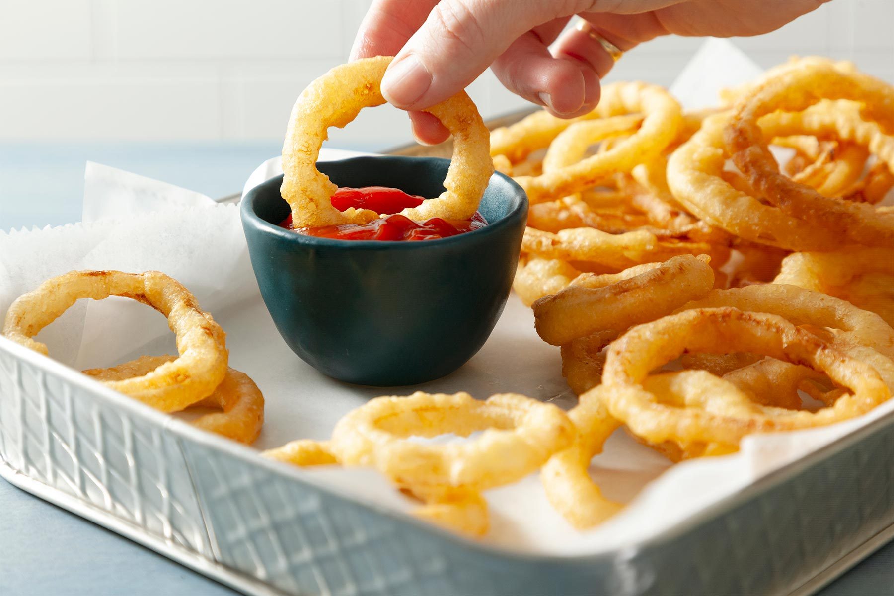 3/4 angle view shot of Dip Crispy Fried Onion Ring in ketchup; served in white tray; light blue background;