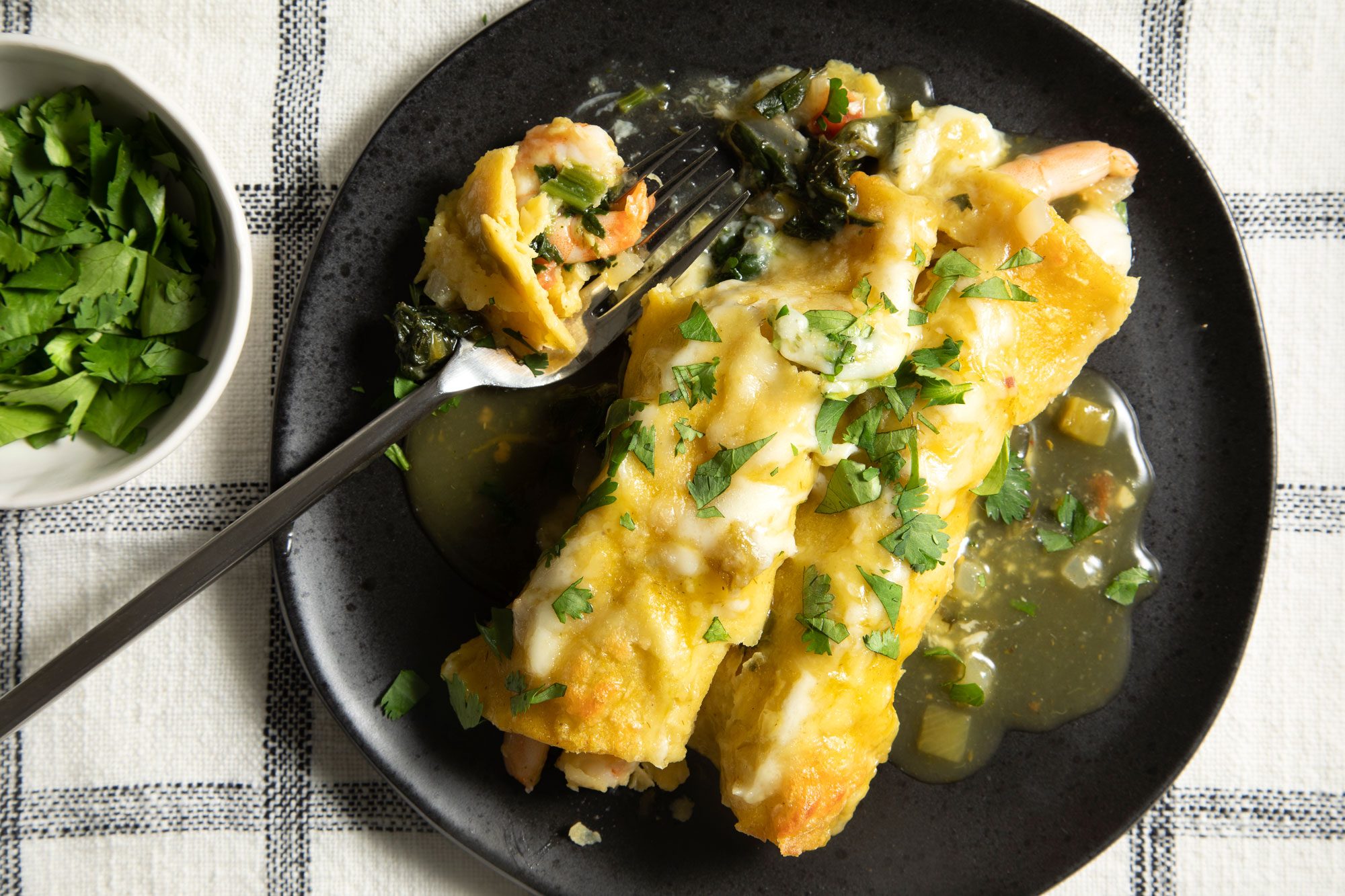 overhead shot; white background; baked Shrimp Enchiladas with Green Sauce served in black plate with silver fork; coriander leaves in small bowl;