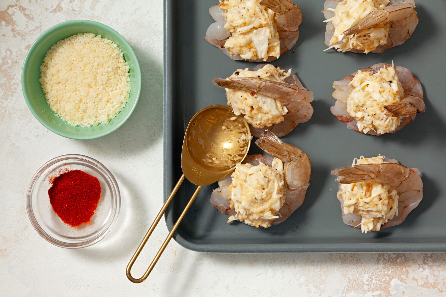 A tray of shrimp prepared with a mixture, next to bowls of cheese and paprika.