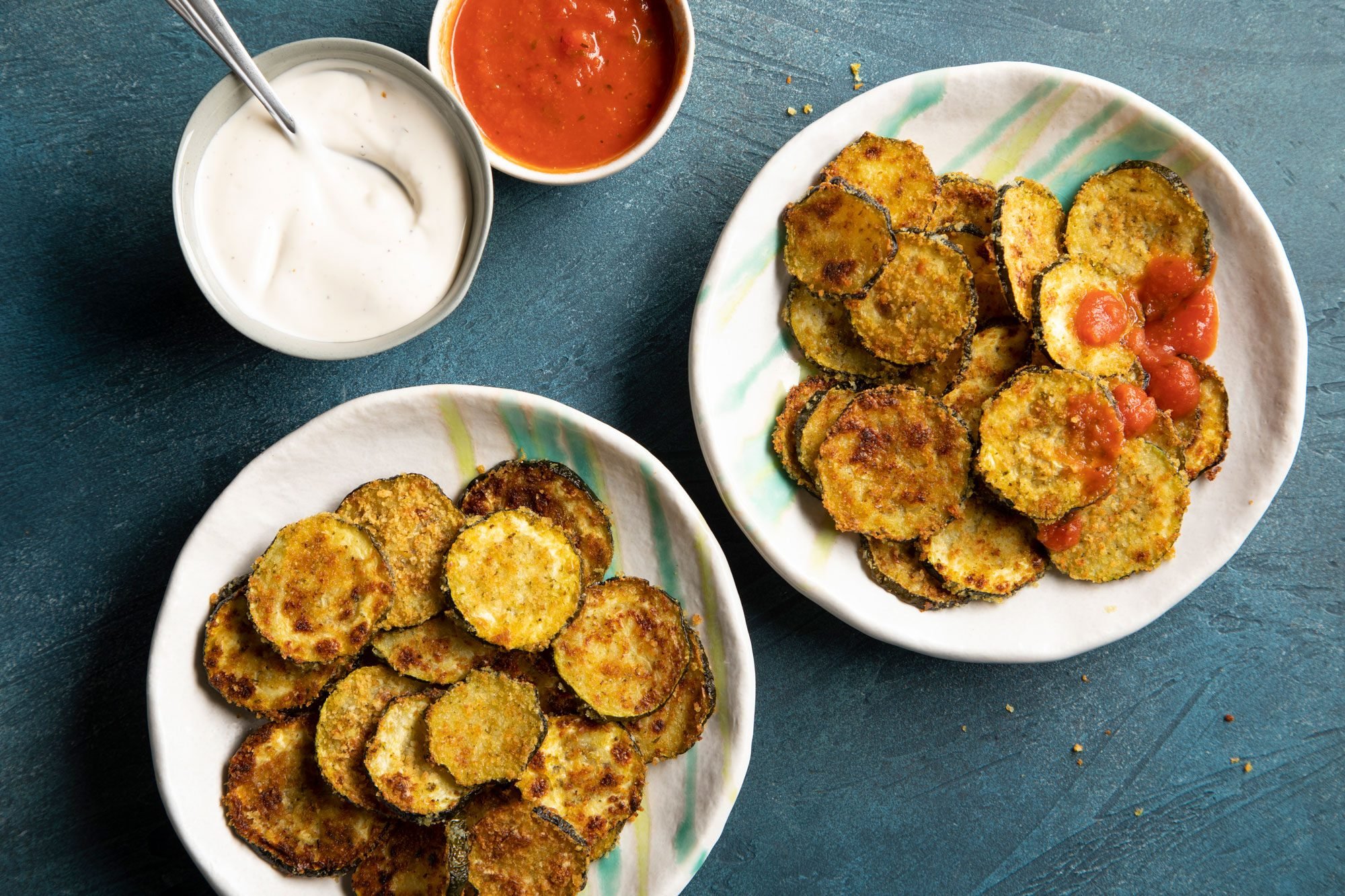 Two bowls of zucchini chips with a side of dipping sauce and parmesan cheese