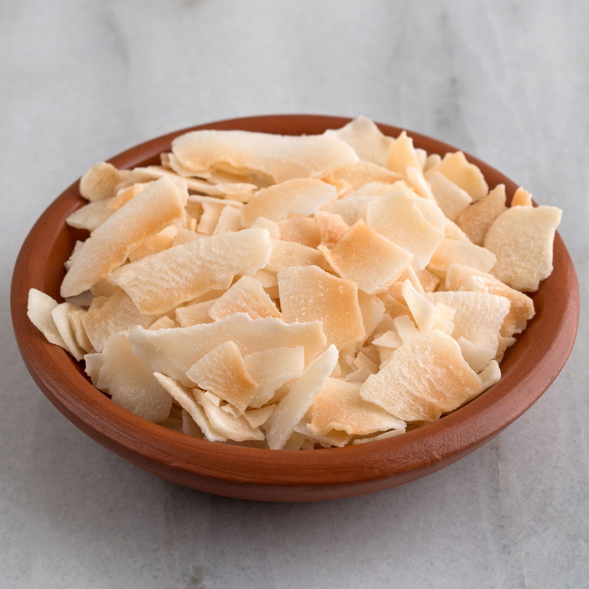 A Small Terracotta Bowl Filled With Toasted Coconut Flakes On A Gray Marble Counter Top