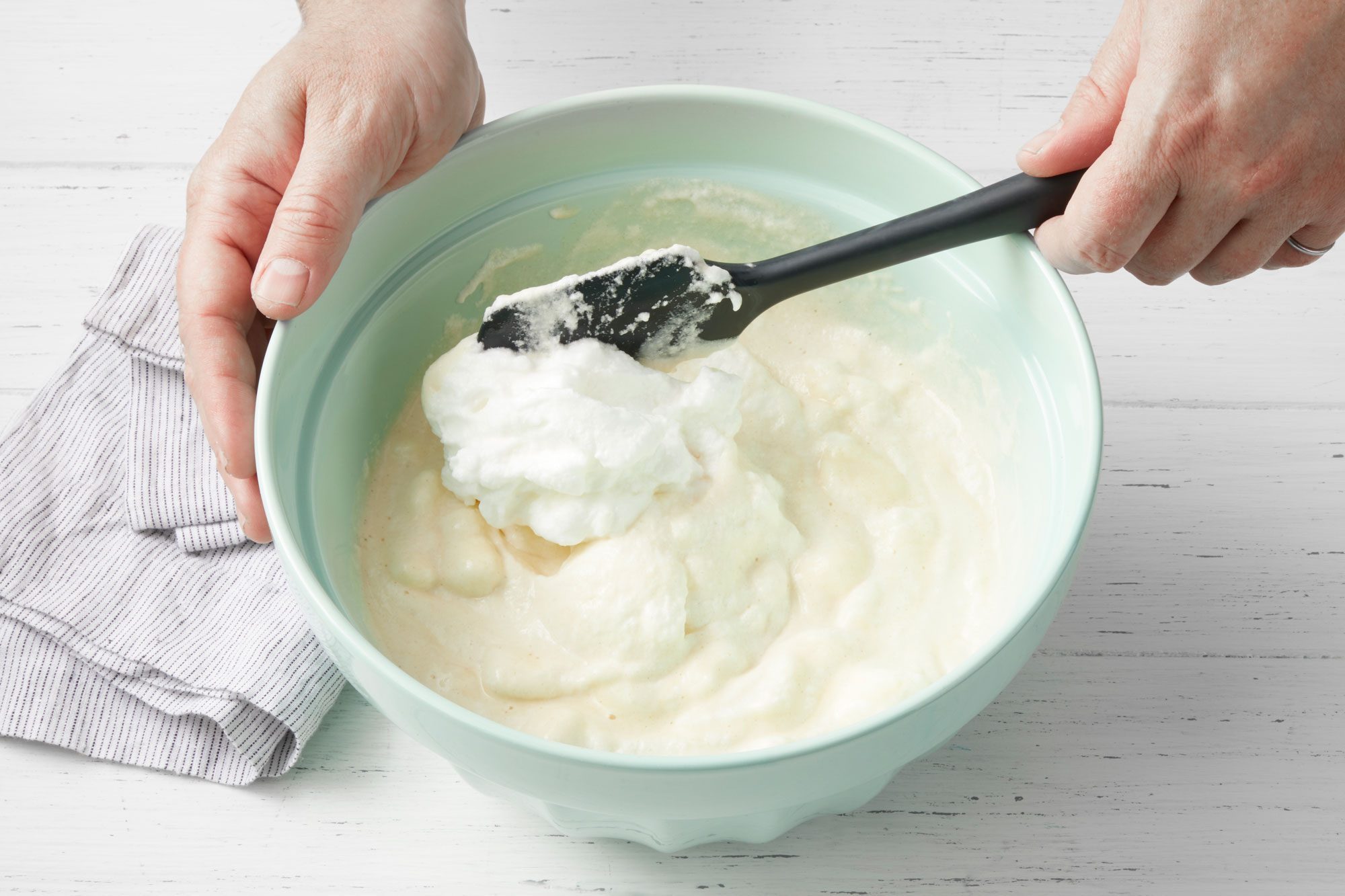 3/4th shot of hands mixing waffles batter in a large mixing bowl using spatula; wooden background;