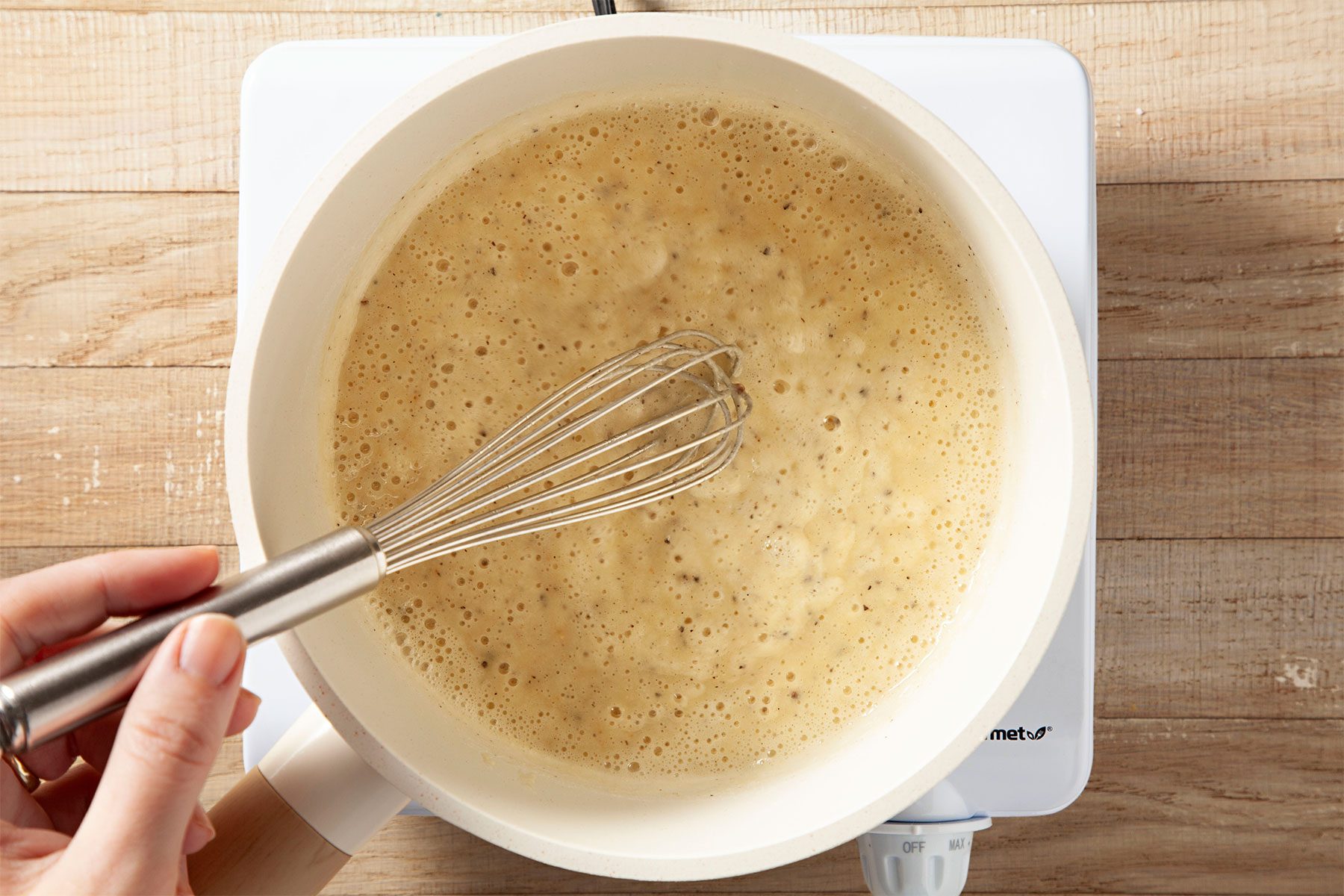 Overhead shot of a small saucepan over medium heat; melt butter; stir in flour and pepper until smooth; whisk tool; induction stove; wooden background;